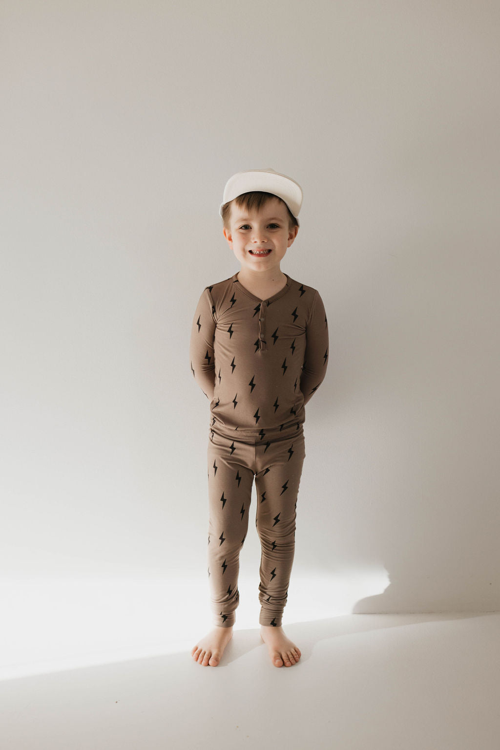A young child stands barefoot against a plain background, wearing the forever french baby Bamboo Two Piece Pajamas in brown with black lightning bolt patterns. The child is smiling and wearing a white hat, hands behind their back.