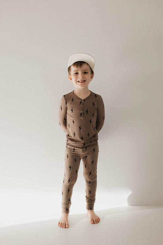 A young boy stands barefoot against a plain background, smiling at the camera. He wears a light brown Bamboo Two Piece Pajamas with a black lightning bolt pattern from forever french baby and accessorizes with a white baseball cap. His hands are behind his back.
