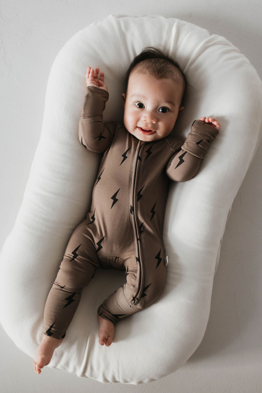 A baby lies on a soft, cushioned white pad, dressed in hypo-allergenic Forever French Baby Bamboo Zip Pajamas. The brown pajamas feature black lightning bolt patterns. With arms raised and a joyful expression, the baby radiates happiness against the plain, light-colored background.