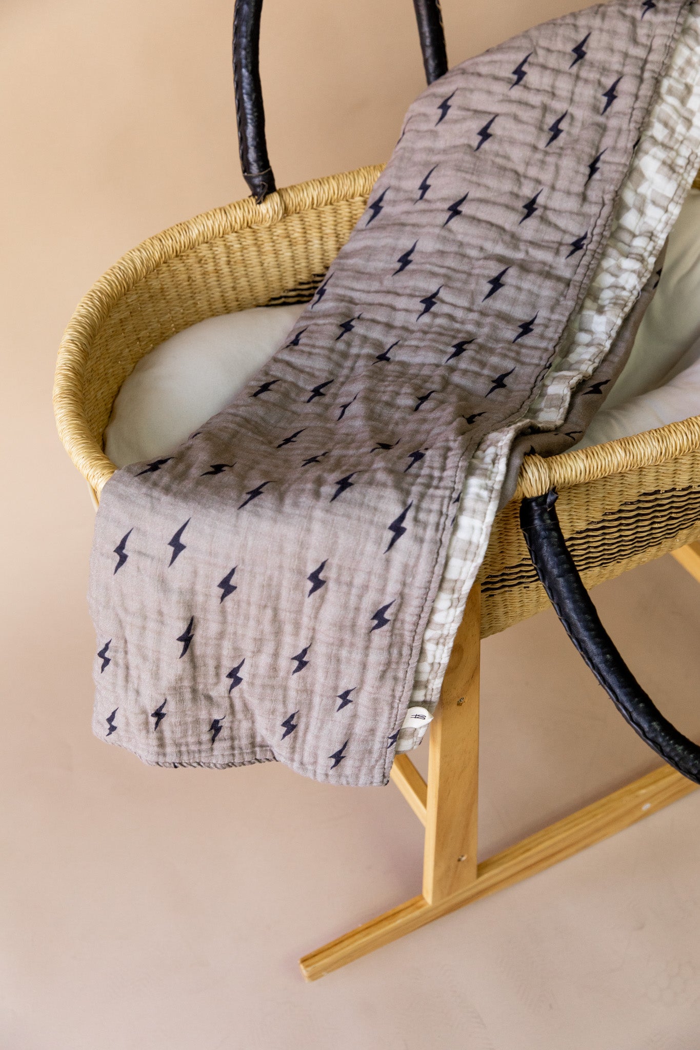 A wicker baby bassinet with black handles and a wooden stand is adorned with the "Muslin Quilt | Lightning Bolt & Wavy Checker" by forever french baby, showcasing light brown muslin cotton featuring black lightning bolt patterns. The 47x47-inch bassinet, complete with a cushion inside, is set against a beige background for an elegant touch from forever french baby.