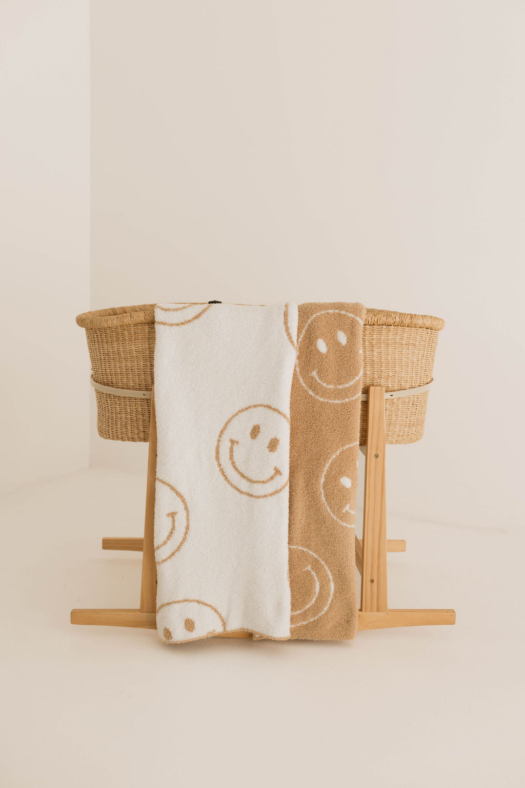 A woven bassinet with wooden legs sits in a minimal room. Two forever french baby Plush Blanket | Original Just Smile, Oeko-tex certified and made from plush microfiber feather yarn, one white with tan smiles and one tan with white smiles, hang over the side.