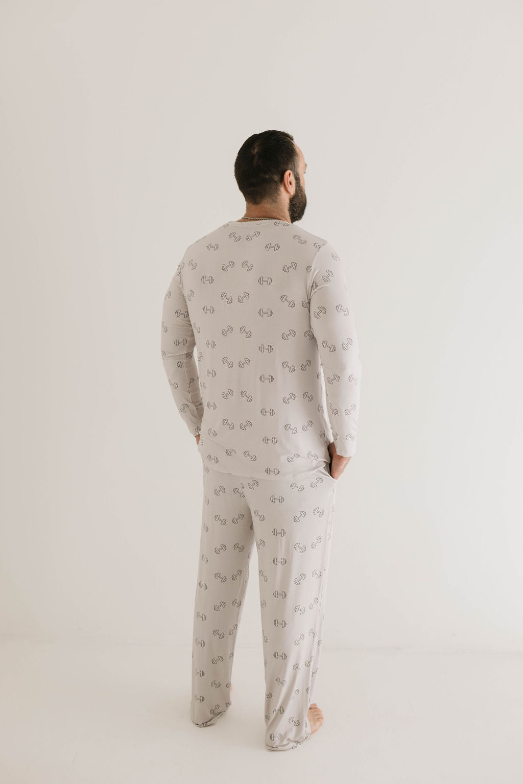A person with short dark hair and a beard stands facing away from the camera, wearing Forever French Baby's Adult Long Sleeve Bamboo Pajamas in the Arm Day design, which features a subtle repeating dumbbell weights graphic. Their hands are tucked into the pockets of the pajama pants. The plain white background suggests a comfortable night’s sleep ahead.