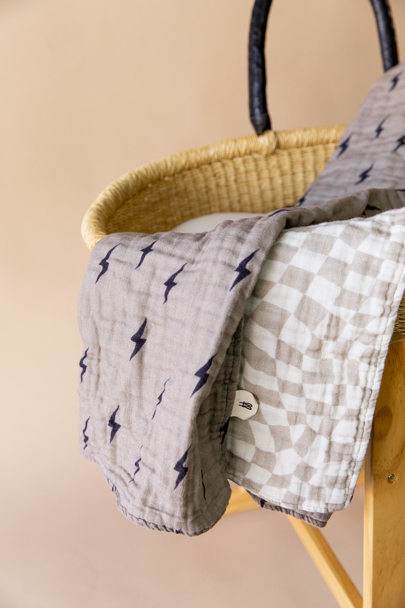 A wicker basket with a dark handle stands on a wooden stand. Two soft blankets from Forever French Baby drape over the edge of the basket – one with a lightning bolt pattern in dark blue on a gray background and the other, featuring a light wavy checker pattern, both part of the Muslin Quilt | Lightning Bolt & Wavy Checker collection. The background is a muted beige.