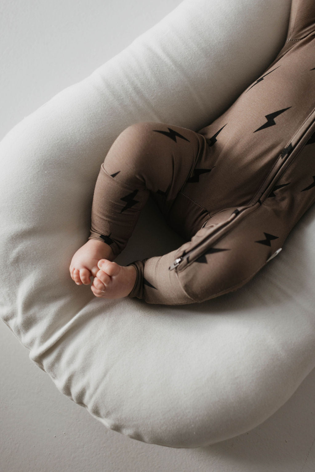 A baby lies on a soft, white cushion, dressed in the Bamboo Zip Pajamas | Brown & Black Lightning Bolt from forever french baby. The image highlights the baby's legs and tiny feet, partially visible against a minimal, light-colored background. The hypo-allergenic outfit promises comfort with its soft premium bamboo fabric.