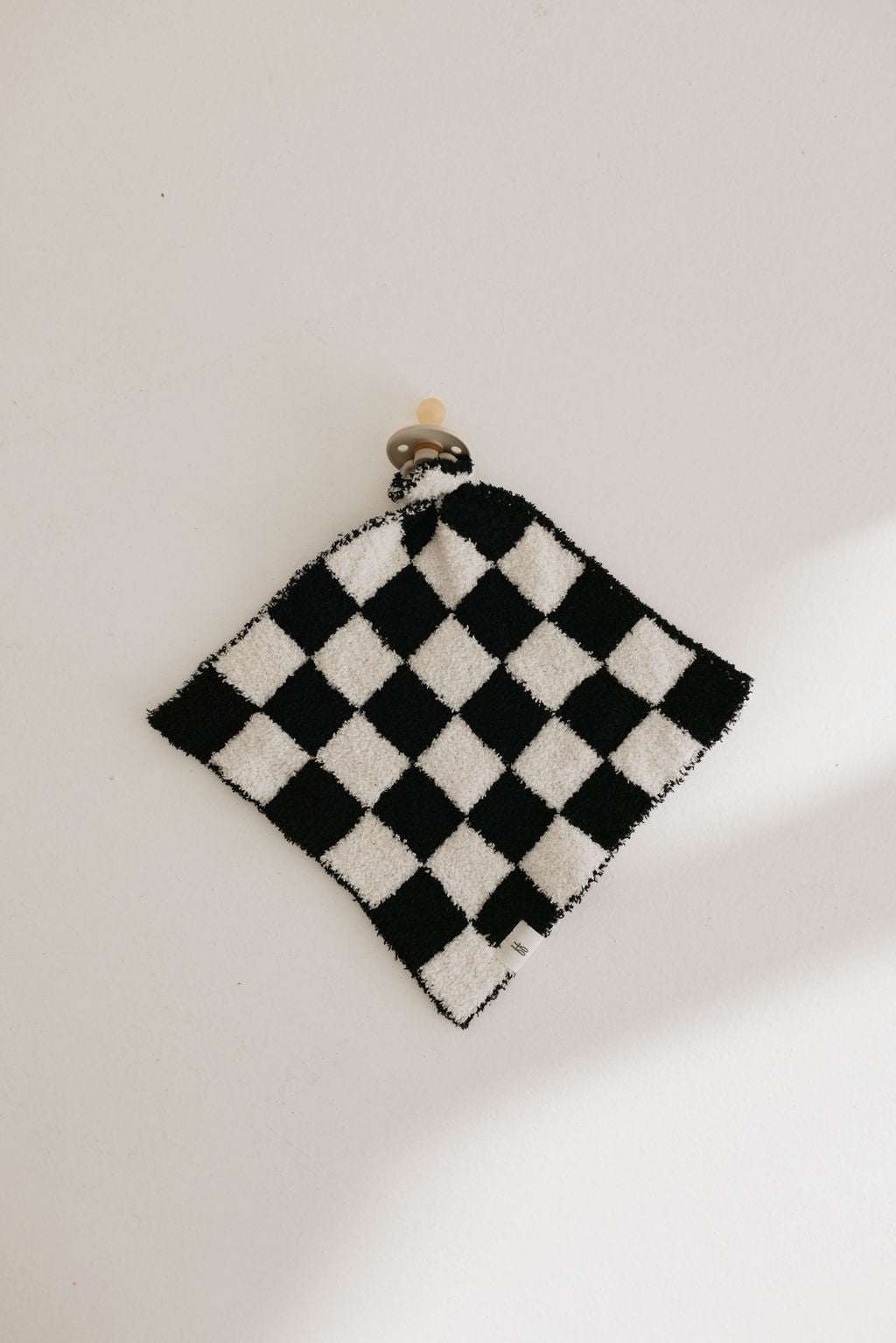 A small Lovey in the Black & White Checkerboard pattern by forever french baby is pinned up against a light-colored wall, secured at the top with a small wooden peg. The microfiber feather yarn gives it a fluffy texture, and there's a visible tag attached to it.
