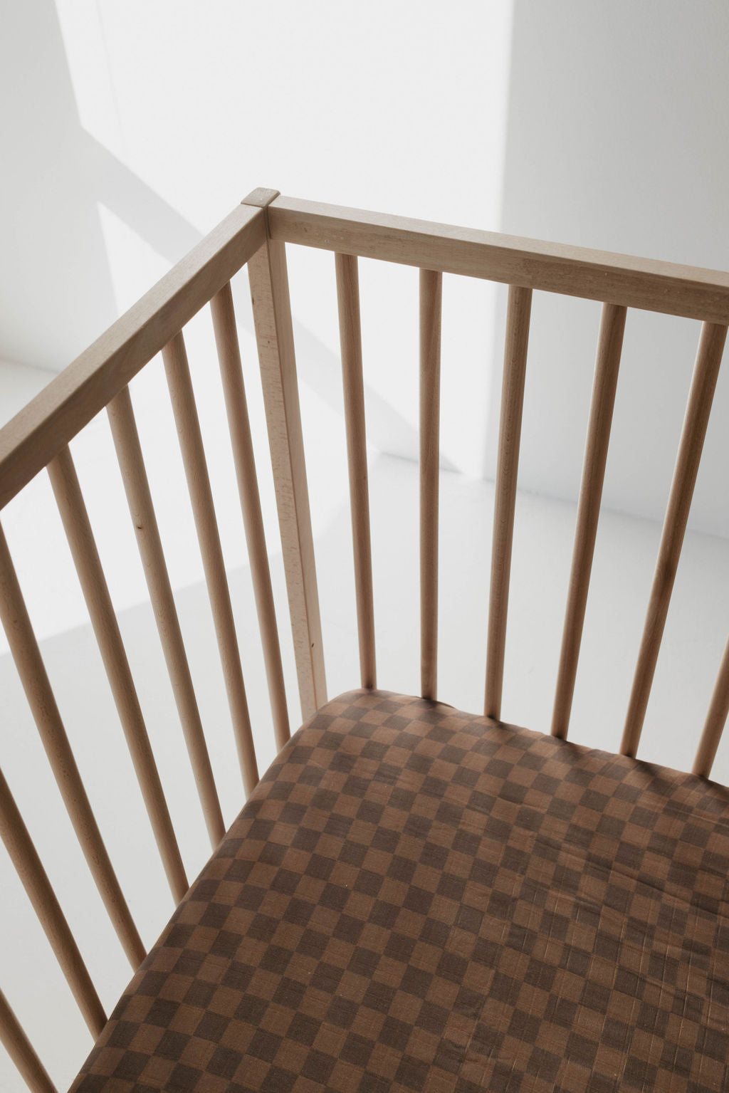 A wooden chair with vertical slats and a brown checkered cushion, similar to the Muslin Crib Sheet | Faded Brown Checkerboard by forever french baby, is bathed in natural light. The sunlight casts soft shadows on the chair, revealing its simple yet elegant design.