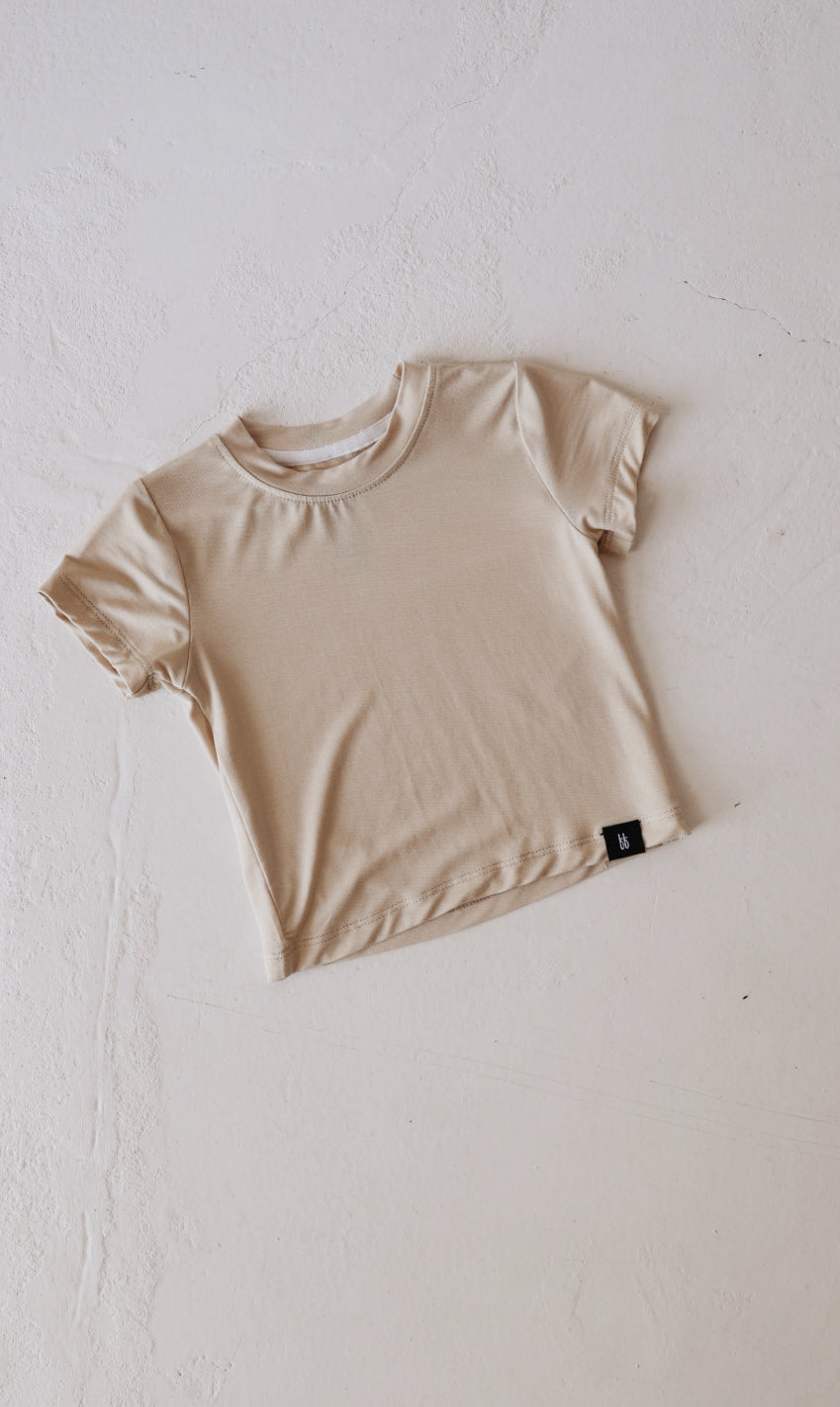 The Bone Basic Bamboo Tee from forever french baby, a beige short-sleeved, plain t-shirt made from soft bamboo fabric, is laid flat on a light, textured surface. The t-shirt has a minimal design with a round neckline and a small black tag on the bottom hem, ensuring comfortable wear for everyday use for babies.