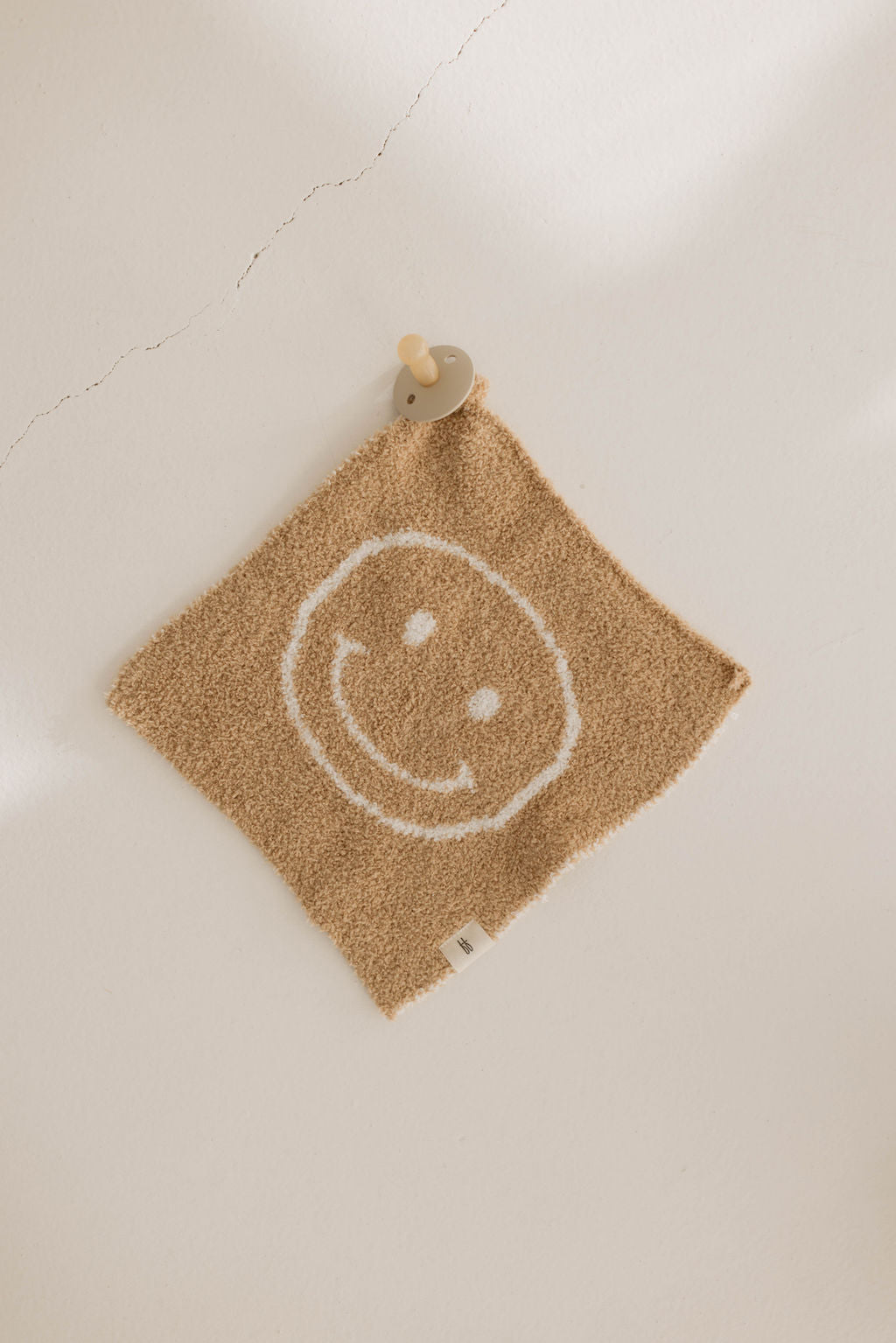 The Lovey | Just Smile Tan washcloth from the forever french baby collection, made from microfiber feather yarn and featuring a white smiley face design, is hanging on a wall with a visible crack running through it. The square beige washcloth is secured using a small wooden hook shaped like a bear’s head.
