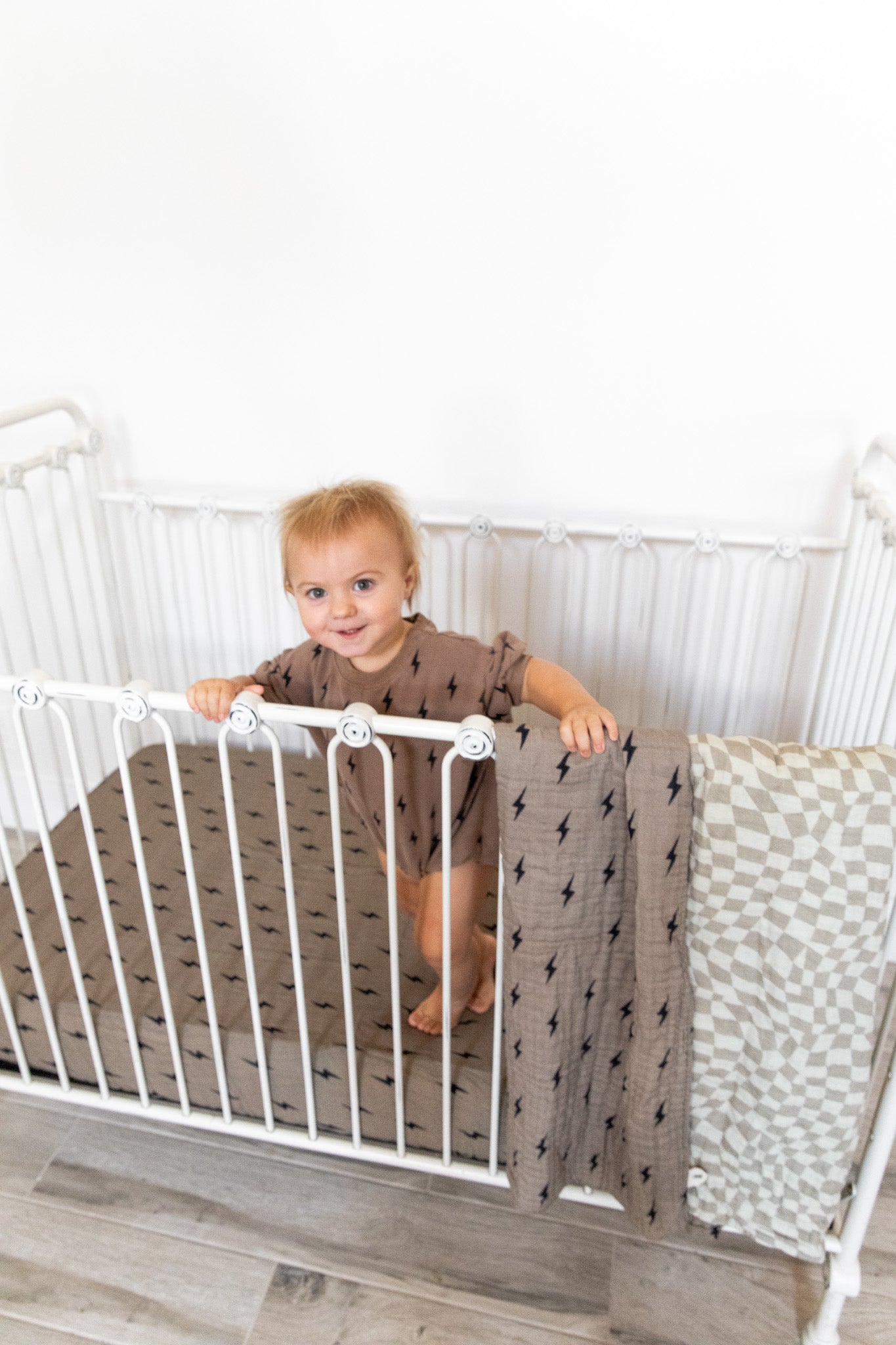 A toddler stands in a white metal crib, holding onto the side rail with both hands. The child wears a brown, short-sleeved onesie with a dark pattern. The crib showcases the Muslin Crib Sheet | Lightning Bolt from forever french baby, and two blankets drape over the side—one muslin cotton with geometric designs and another adorned with lightning bolts. The room features light-colored flooring and a white wall.