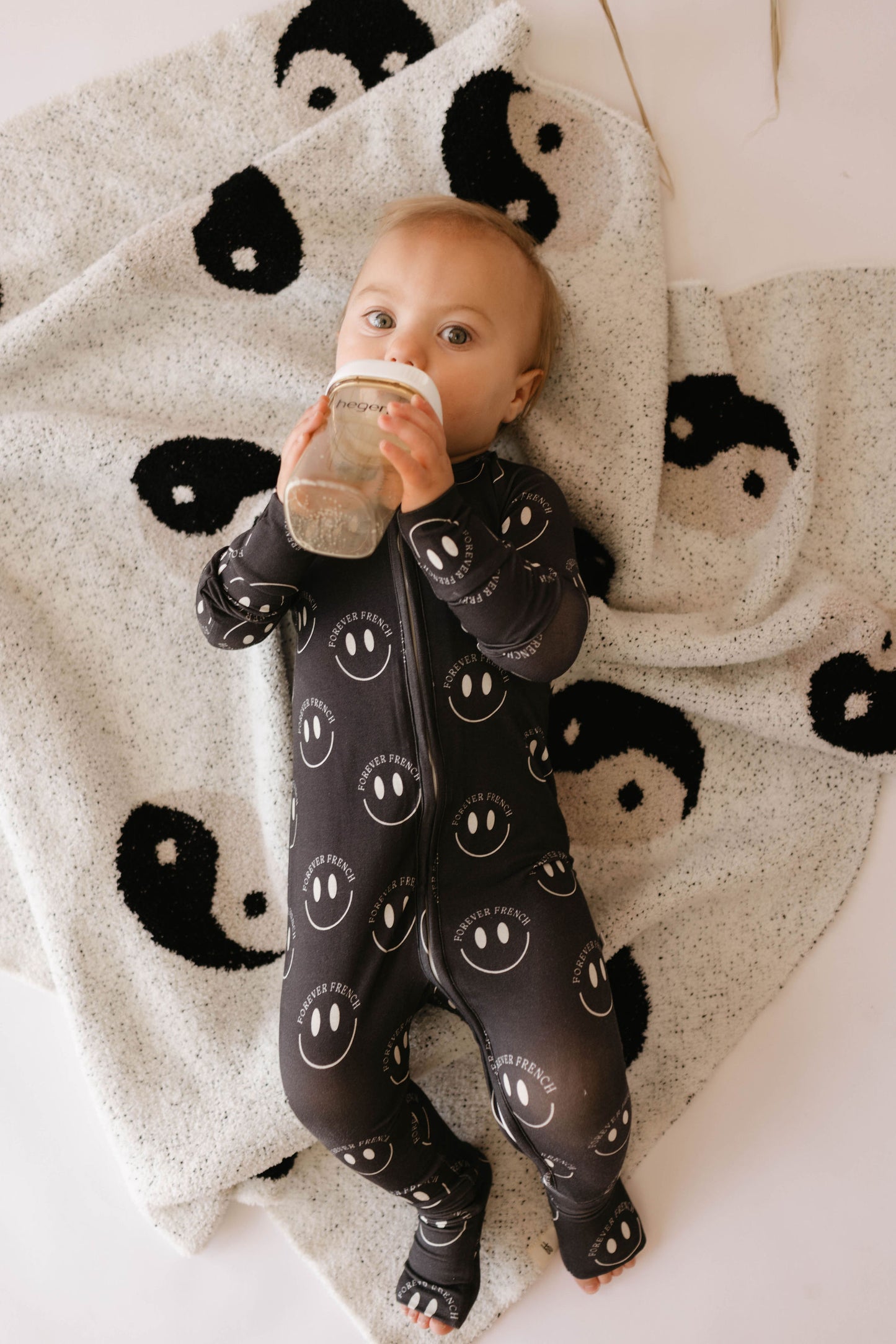 A baby in a black onesie with white smiley faces lays on a forever french baby's Plush Blanket | Black & White Yin Yang. The velvety-rich texture of the microfiber feather yarn ensures comfort, as the baby holds a bottle and looks up contentedly.