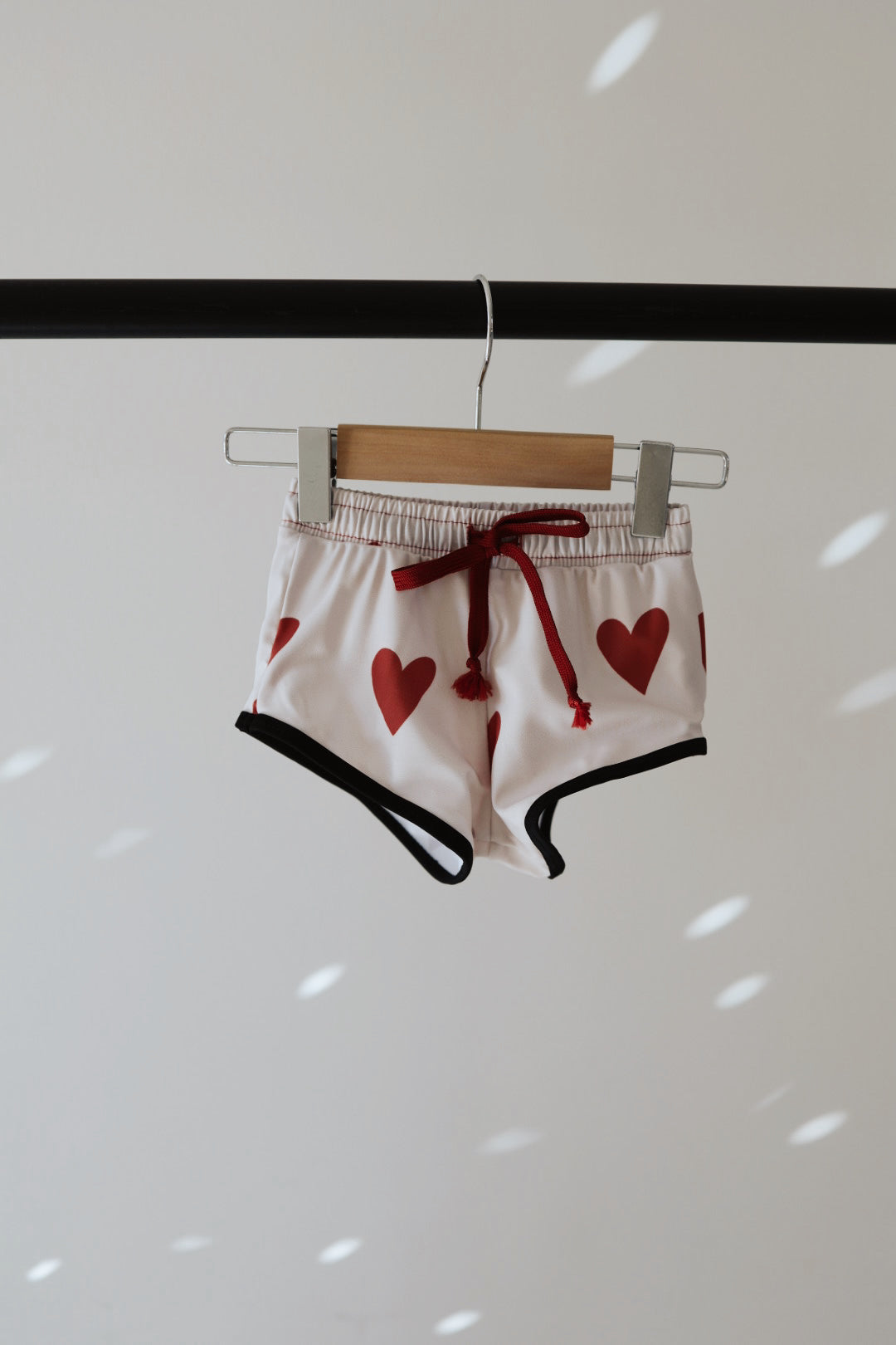The King of Hearts Boys Board Short by forever french baby, featuring white fabric with red heart prints and black trim, is hanging on a wooden hanger against a plain background. Perfect for swimwear, these shorts have an elastic waistband with a red drawstring. Soft, diffused light casts gentle spots on the wall behind.