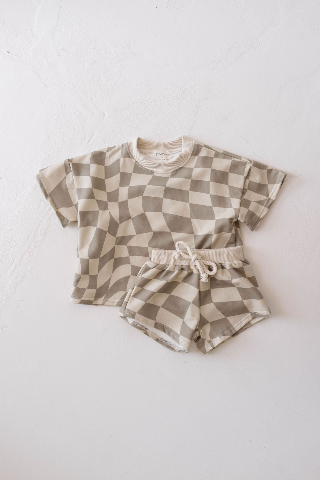 A Short Set | Wavy Checkerboard by forever french baby, featuring a beige and white optical illusion pattern, lies flat on a white surface. Crafted from organic cotton, the trendy ensemble includes a short-sleeve T-shirt with a rounded neckline and matching shorts with a drawstring waist.