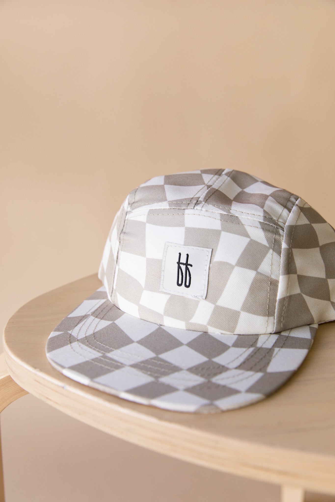 A chic Children's Snap | Wavy Checker five-panel hat from forever french baby, featuring a stylish beige and white checkerboard pattern, rests on a wooden stool. The hat includes a white rectangular patch on the front with a black double "b" logo. Suitable for ages 4 months to 6 years, this accessory is easy to spot clean.