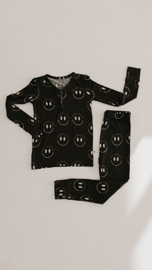 A relaxed set of Bamboo Two Piece Pajamas in Charcoal & White ff Smile from forever french baby is arranged on a light-colored surface. Made from breathable, hypo-allergenic fabric, the ensemble includes a top and matching pants, both adorned with the same whimsical white smiley face pattern.