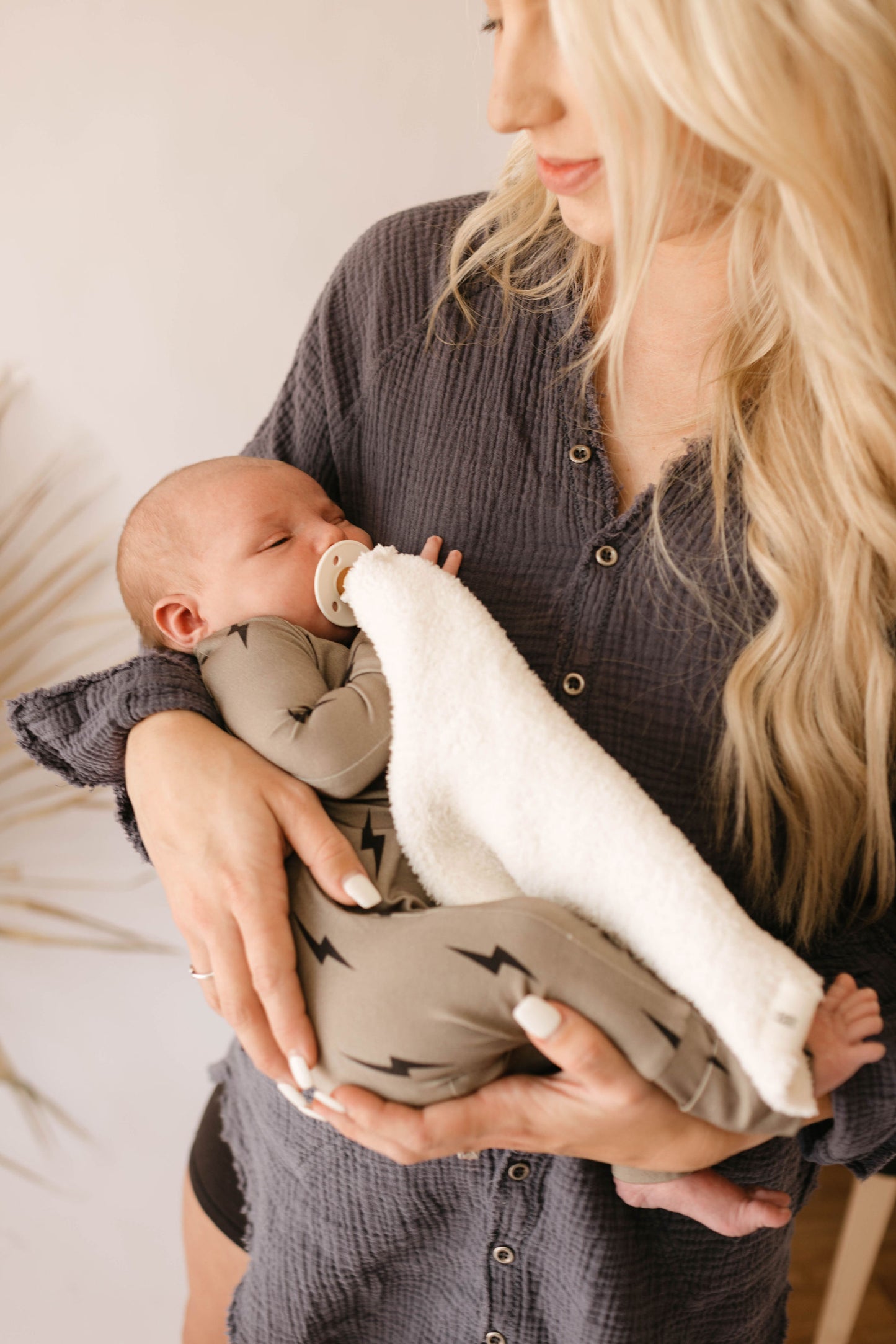 A woman with long blonde hair, wearing a dark button-up shirt, gently cradles a sleeping baby dressed in Forever French Baby Bamboo Zip Pajamas with brown and black lightning bolt patterns. The baby is covered with a white hypo-allergenic blanket and has a pacifier in their mouth.