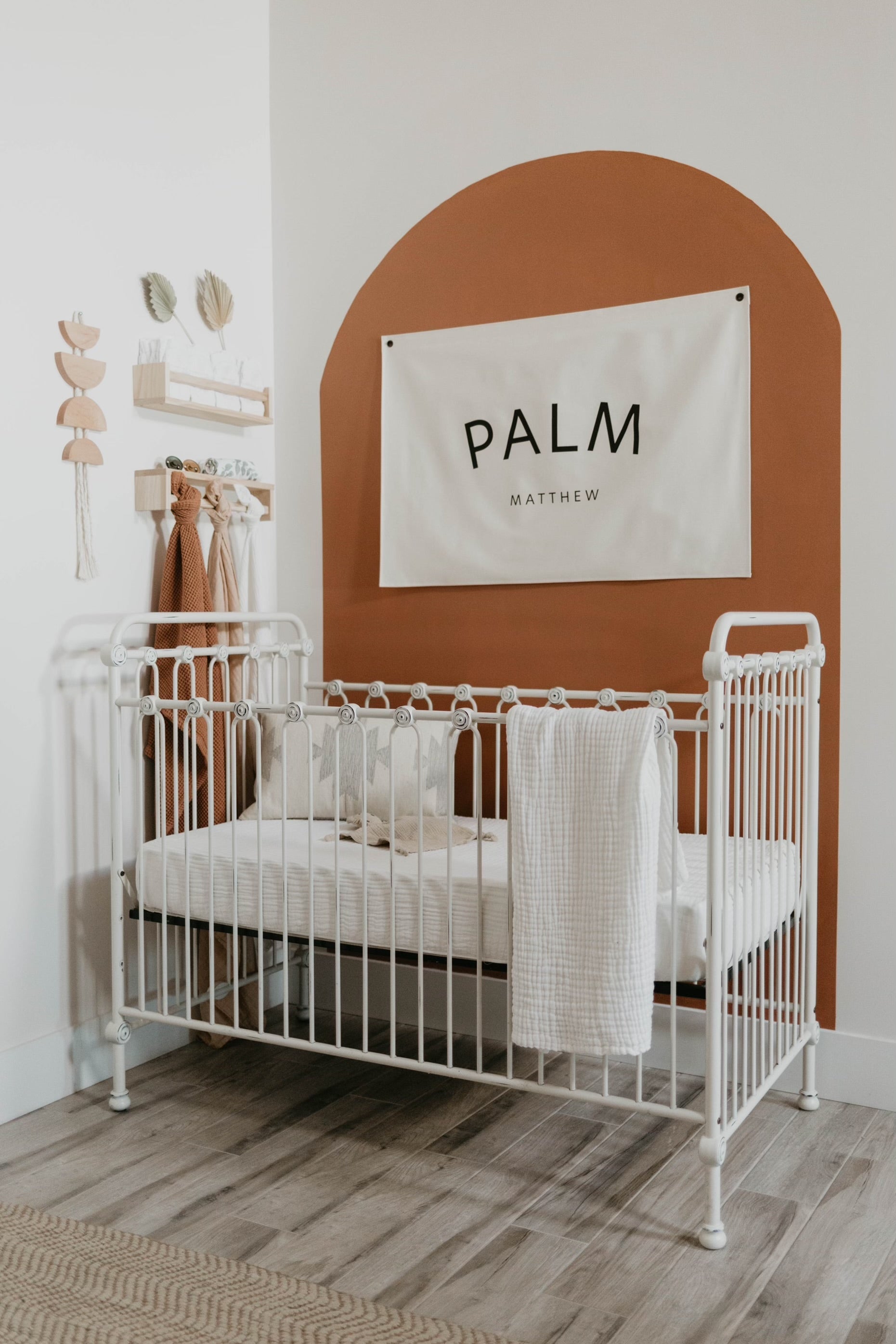 A minimalistic nursery features a white metal crib against a white wall with a rust-orange arch accent. Above the crib hangs a banner with the text "PALM Matthew." The room includes wooden floors, light-colored decor items, and features the charming Quilt | Cloud from Forever French Baby as part of its decor.