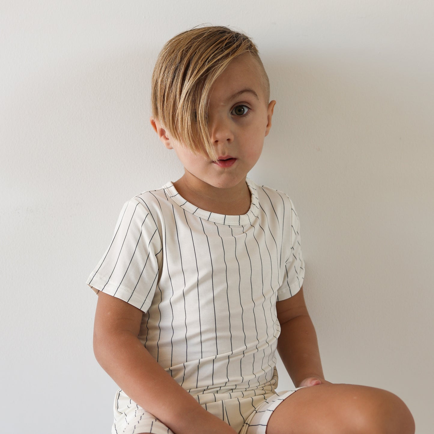 A young child with light brown side-swept hair wears forever french baby's Bamboo Short Set | Jack—a breathable white shirt with thin black stripes—while sitting against a plain white background and looking surprised.