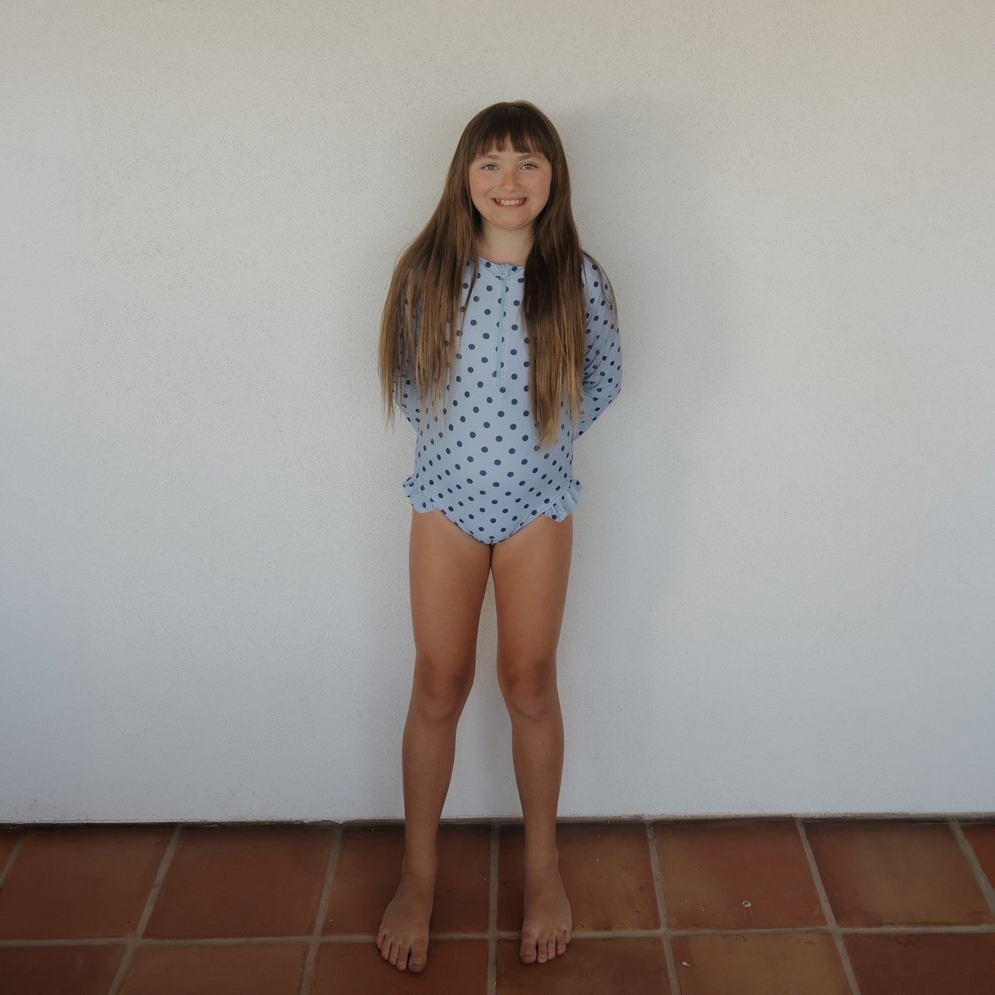 A young girl with long brown hair smiles at the camera, barefoot on a tiled floor. She wears the forever french baby Girl's Long Sleeve Swimsuit | Sully in light blue with dark polka dots. The wall behind her is plain white.