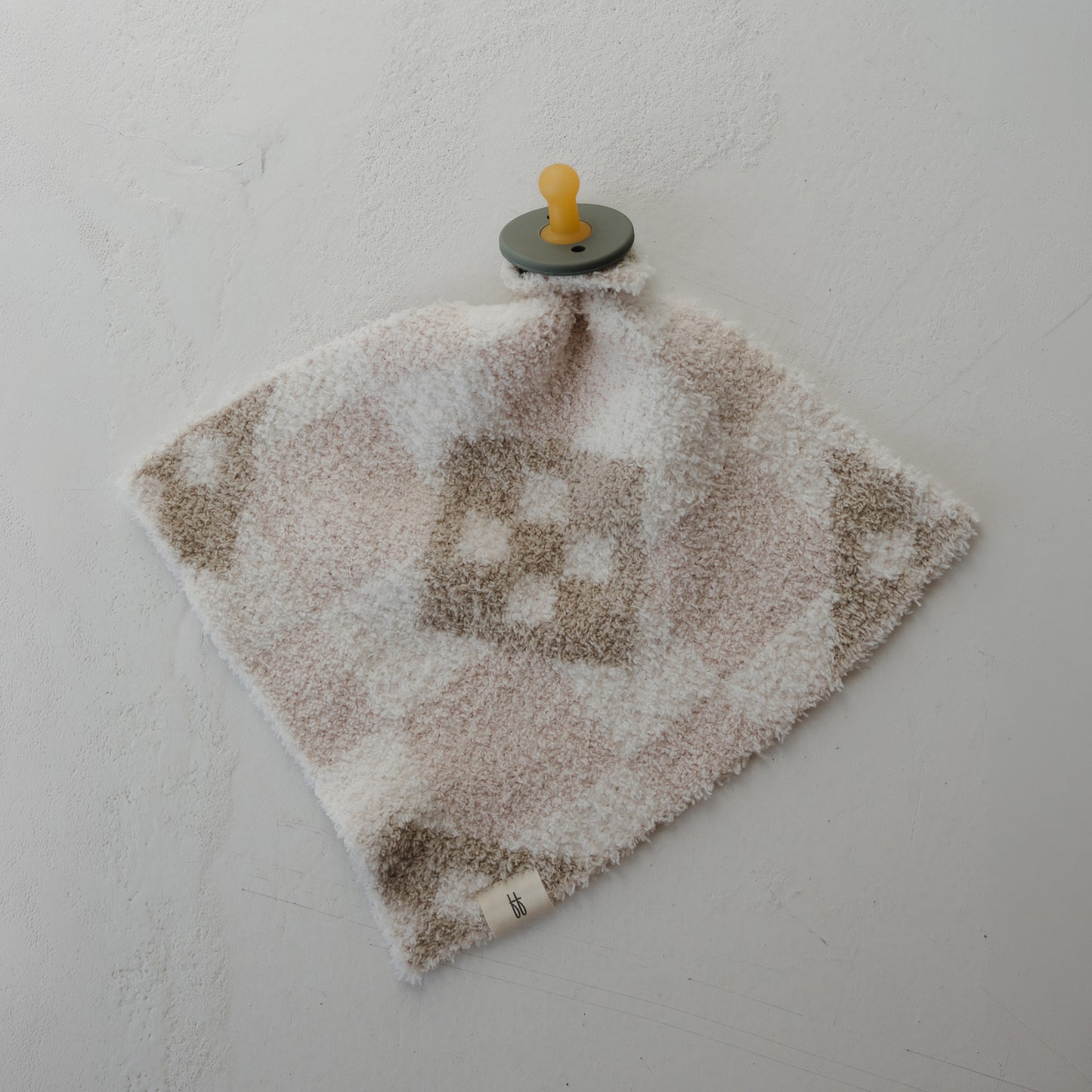 The Lovey | Cottage Quilt by forever french baby is a soft, square comfort blanket with a beige and white check pattern, made from cozy microfiber feather yarn, featuring an attached pacifier and shown on a light textured surface.