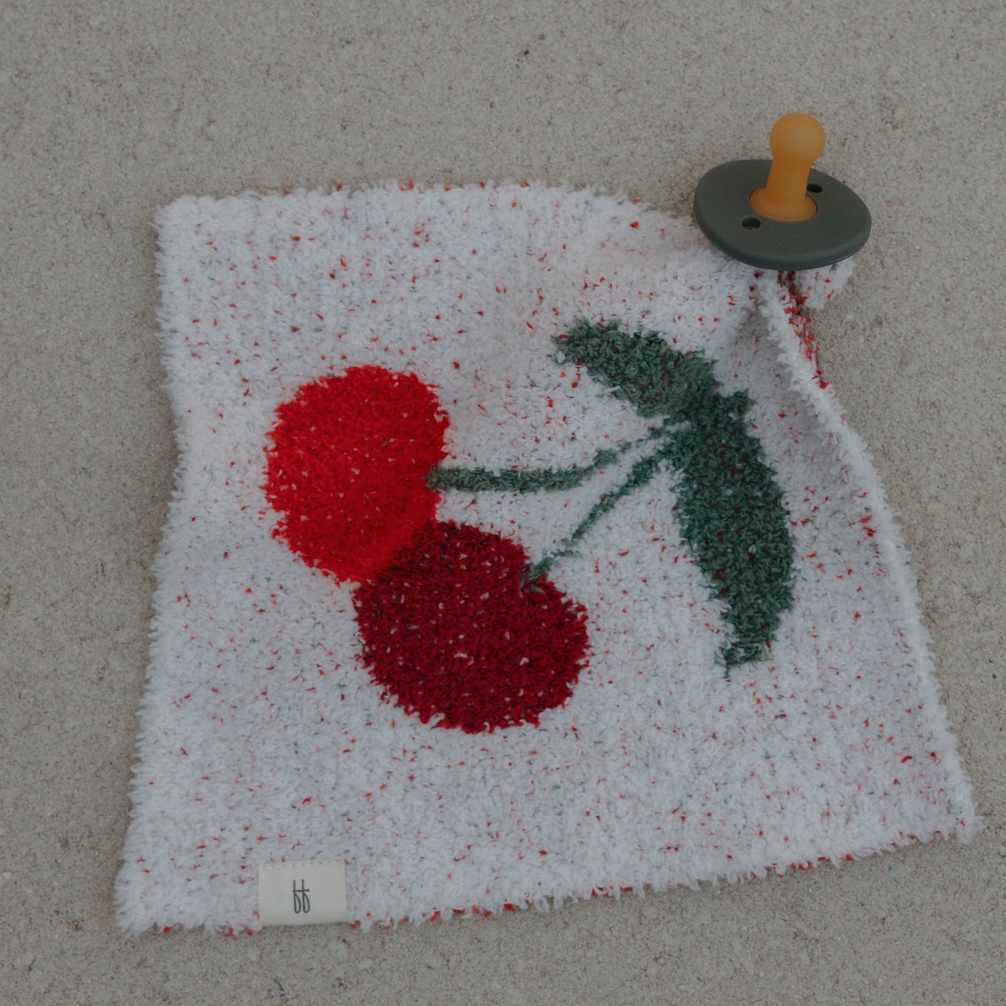 The forever french baby Lovey | Cherry is a soft, white microfiber security blanket with a cherry print and an attached pacifier, featuring a gray base and tan nipple, shown lying on a light surface.
