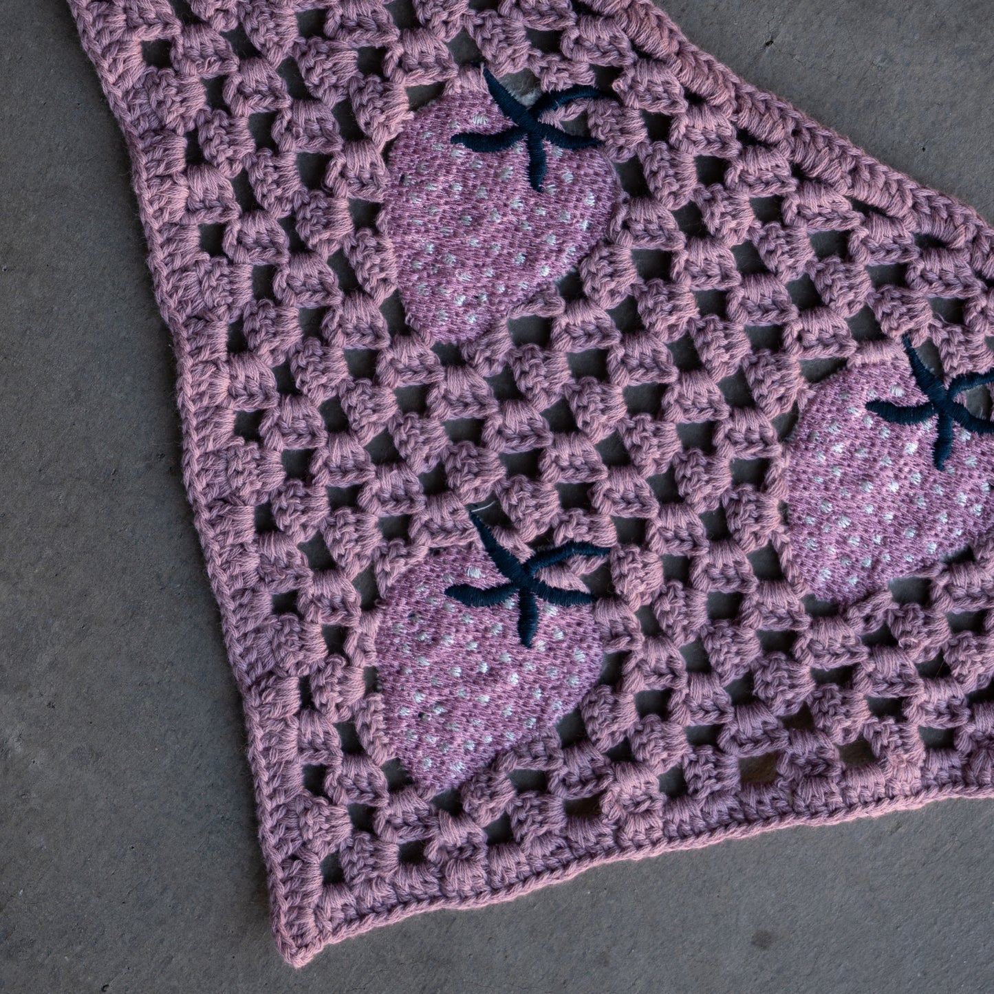 The Knit Head Scarf | Matilda by forever french baby is a triangular crochet accessory crafted from soft cotton yarn, featuring a purple border and adorned with multiple pink strawberry motifs topped with green. Designed for versatility and comfort, it includes an adjustable tie for the perfect fit, and is shown here laid on a gray surface.