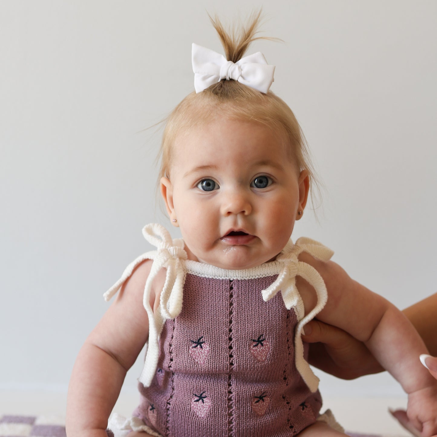A light-haired, blue-eyed baby wears the forever french baby Knit Ruffle Romper | Matilda in pink with white bows and a matching hair bow, sitting upright with gentle support against a plain, light background.