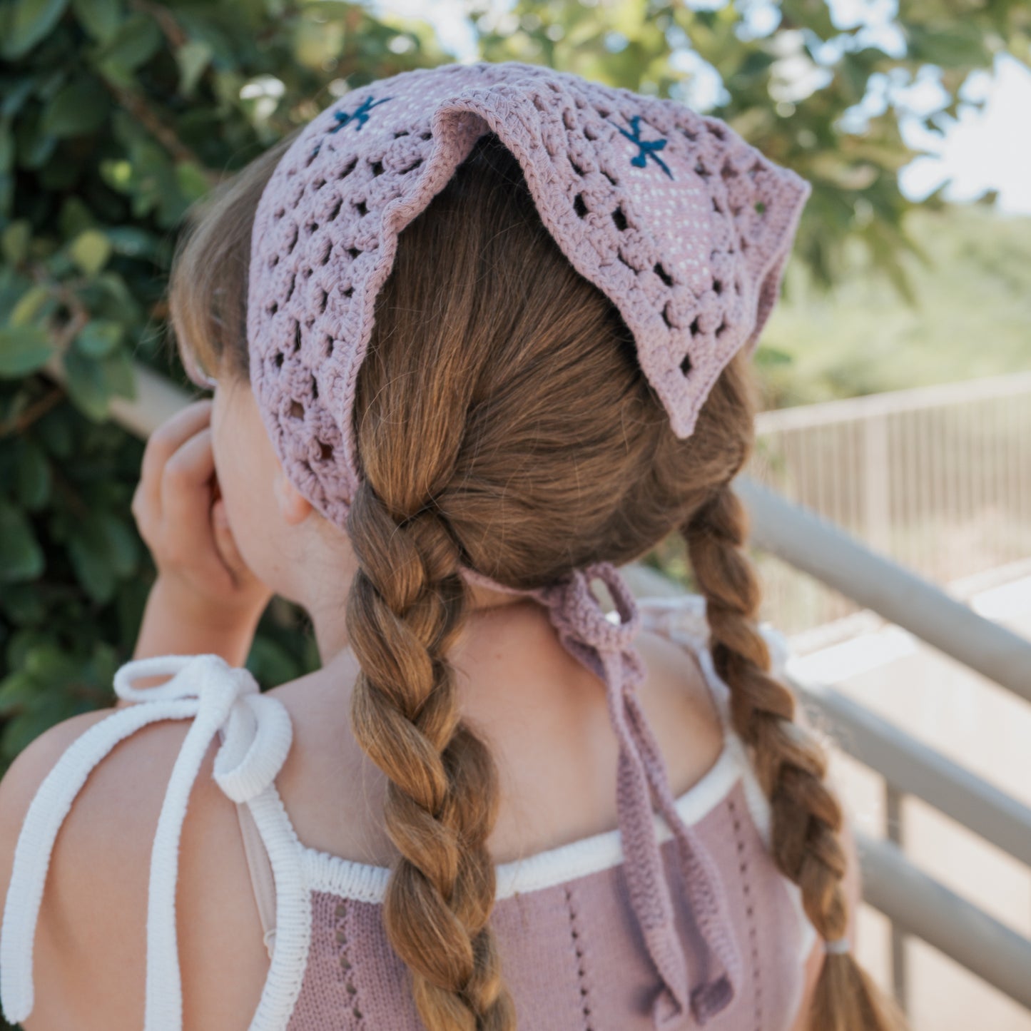 A young girl with two braids wears the forever french baby Knit Dress | Matilda, a sleeveless 100% cotton dress with adjustable tie straps. She stands outdoors near a railing, facing away from the camera, in a light purple crocheted headscarf.