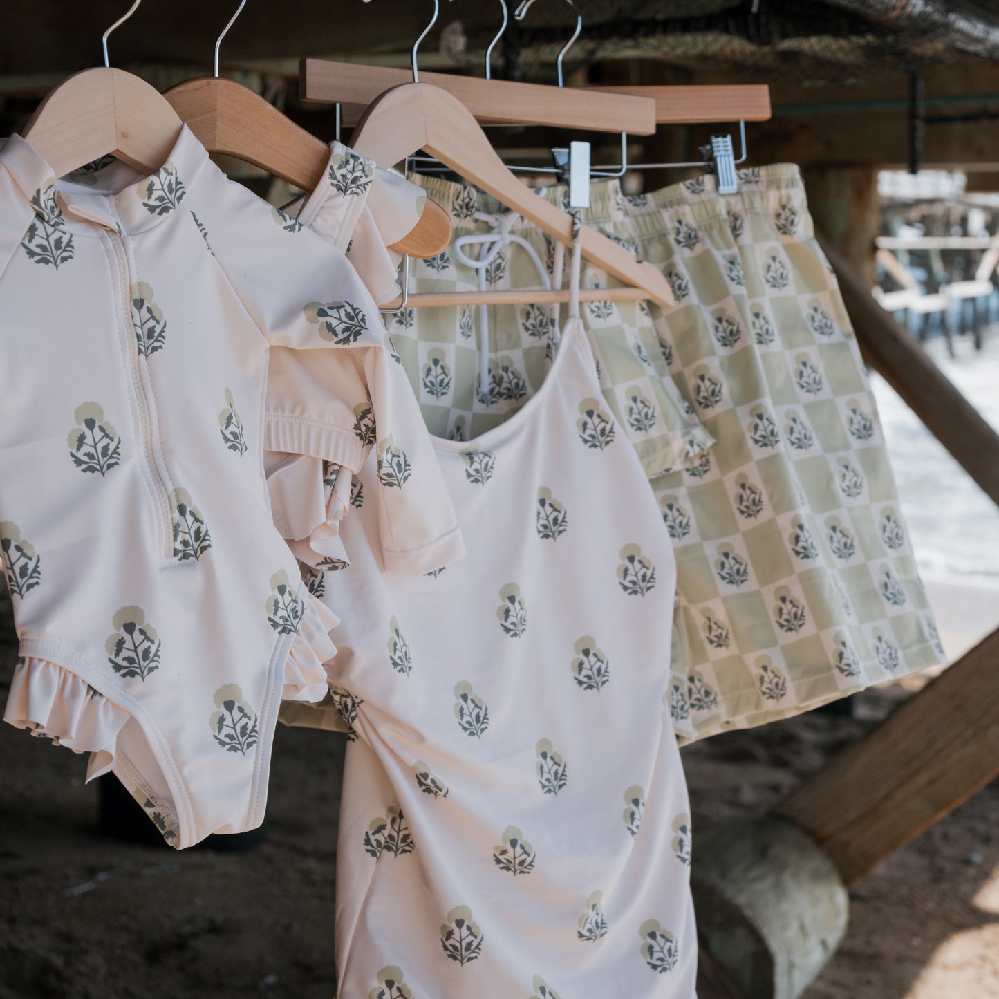 A light floral Girl's Two Piece Swimsuit | Marigold by forever french baby hangs on a wooden hanger, with sand and wooden beams in the background—perfect beach day vibes for lovers of stylish kids swimwear.