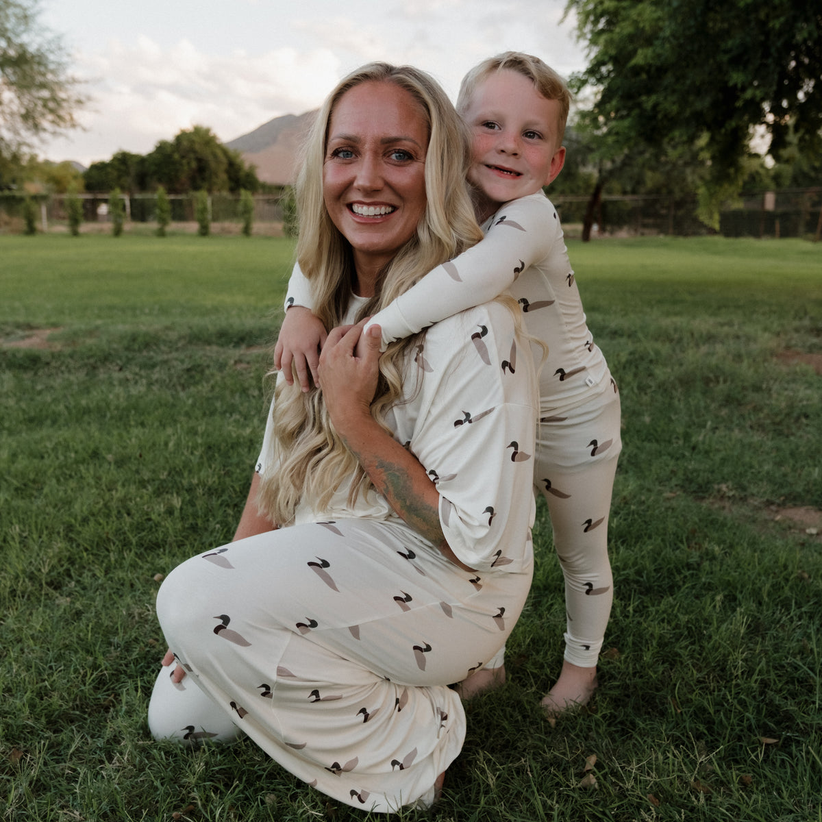 A woman kneels on the grass, smiling, as a young child in matching forever french baby Bamboo Two Piece Pajamas | Mallard hugs her from behind. Trees and a mountain are visible in the background.