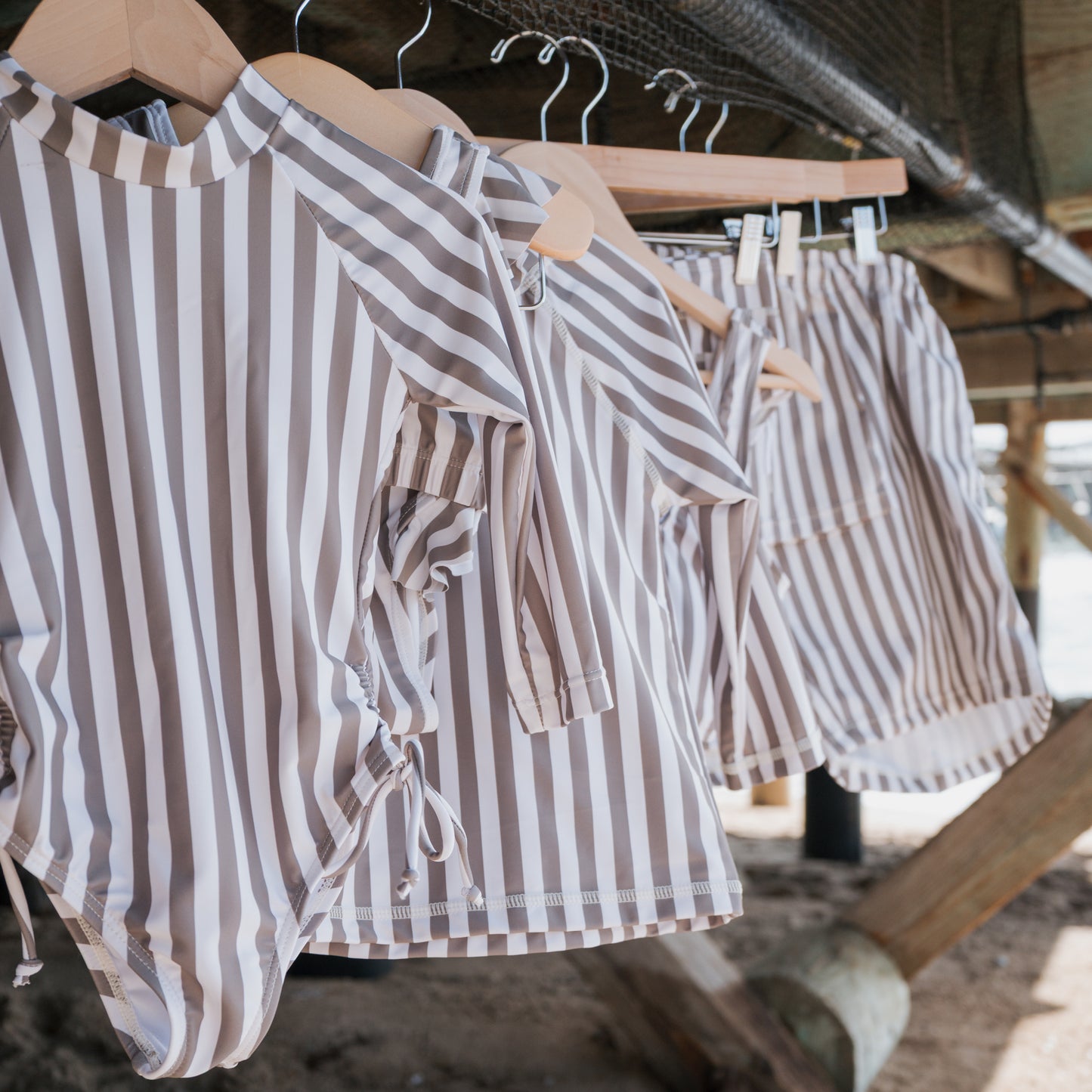Forever french baby Girl's Two Piece Swimsuit in Olive Stripe hangs on a wooden hanger under a wooden structure, with sand and wooden beams seen in the background.