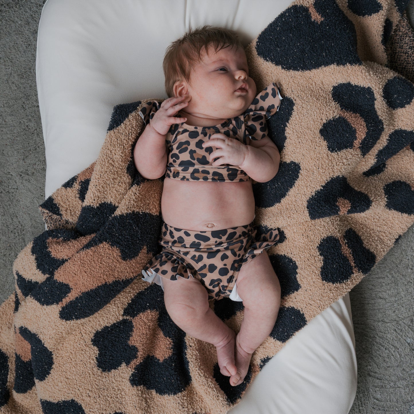 A baby with light brown hair lies on a white cushion, looking up and wearing the forever french baby *PRE-ORDER* Girl's Two Piece Swimsuit | Leopard, paired with a brown and black leopard print blanket.
