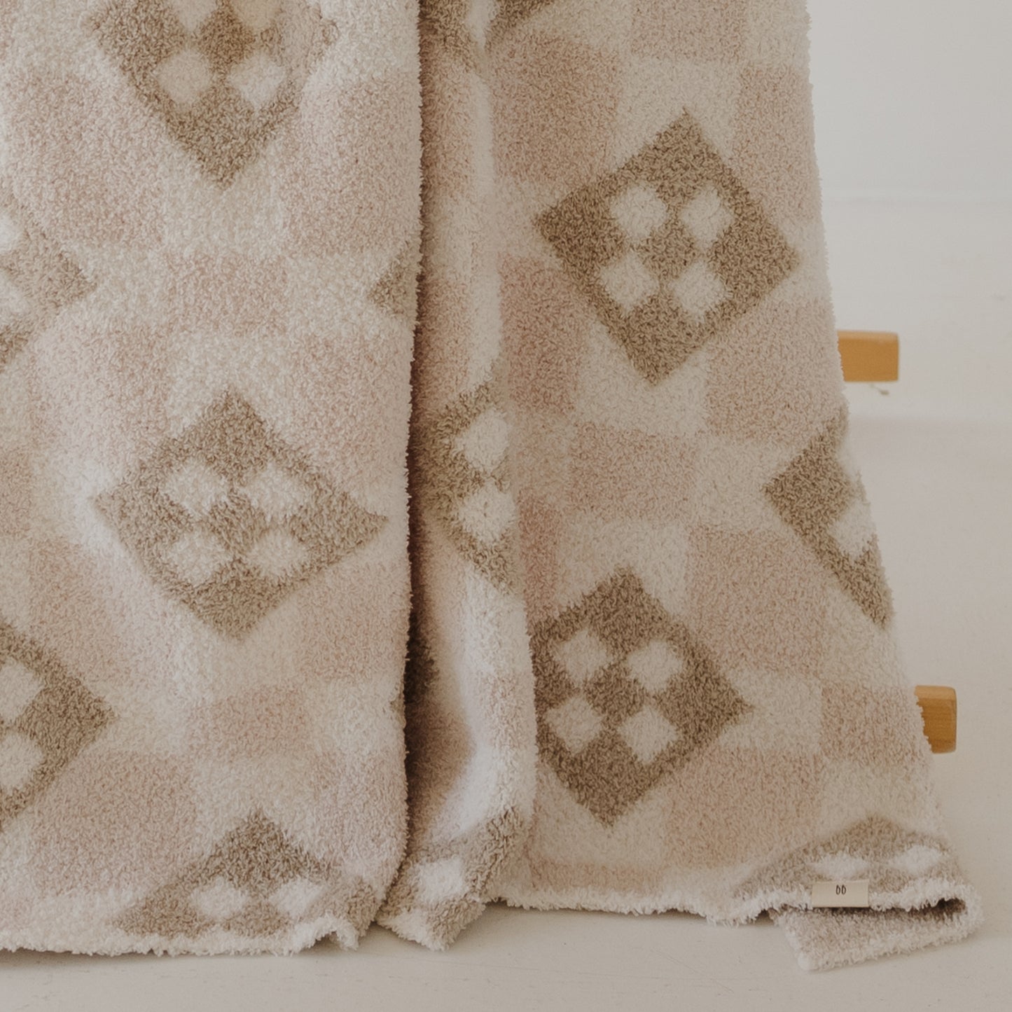 The forever french baby Plush Blanket | Cottage Quilt in beige and cream, with a geometric diamond pattern, drapes over a light wooden stand and partially touches the white floor.