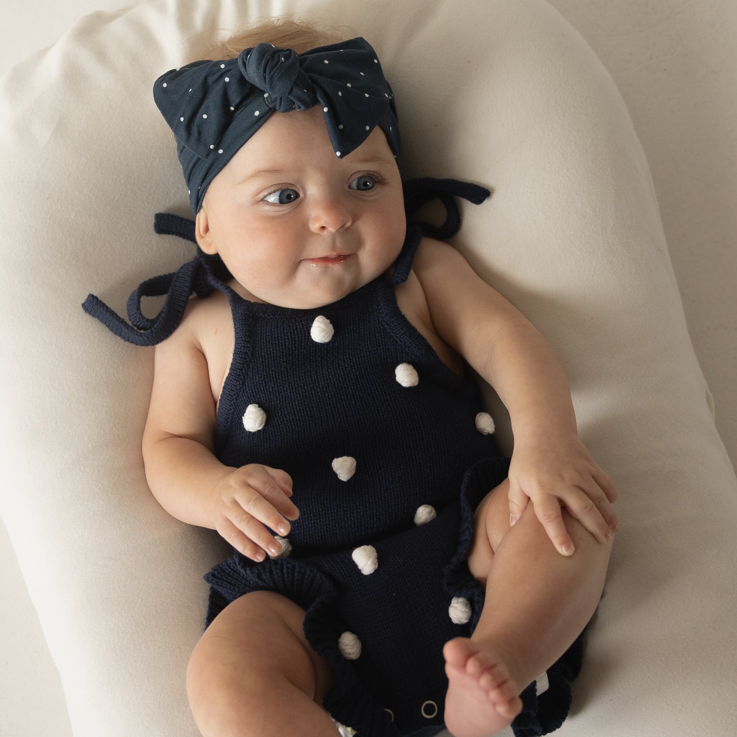 A baby in a forever french baby navy outfit with white pom-poms and wearing the Bamboo Head Wrap | Emery sits on a white cushion, looking slightly to the side.
