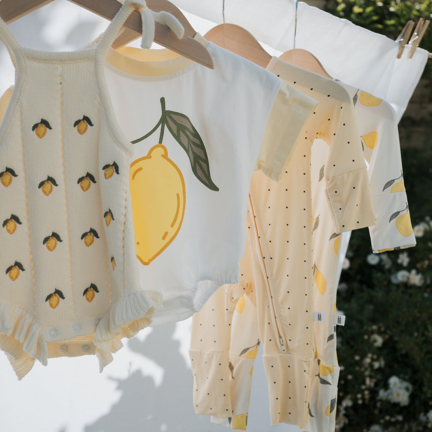 Four baby outfits, including the forever french baby Short Sleeve Romper in Lemon, hang on wooden hangers outdoors amid sunlight and greenery—perfect inspiration for a playful summer look.