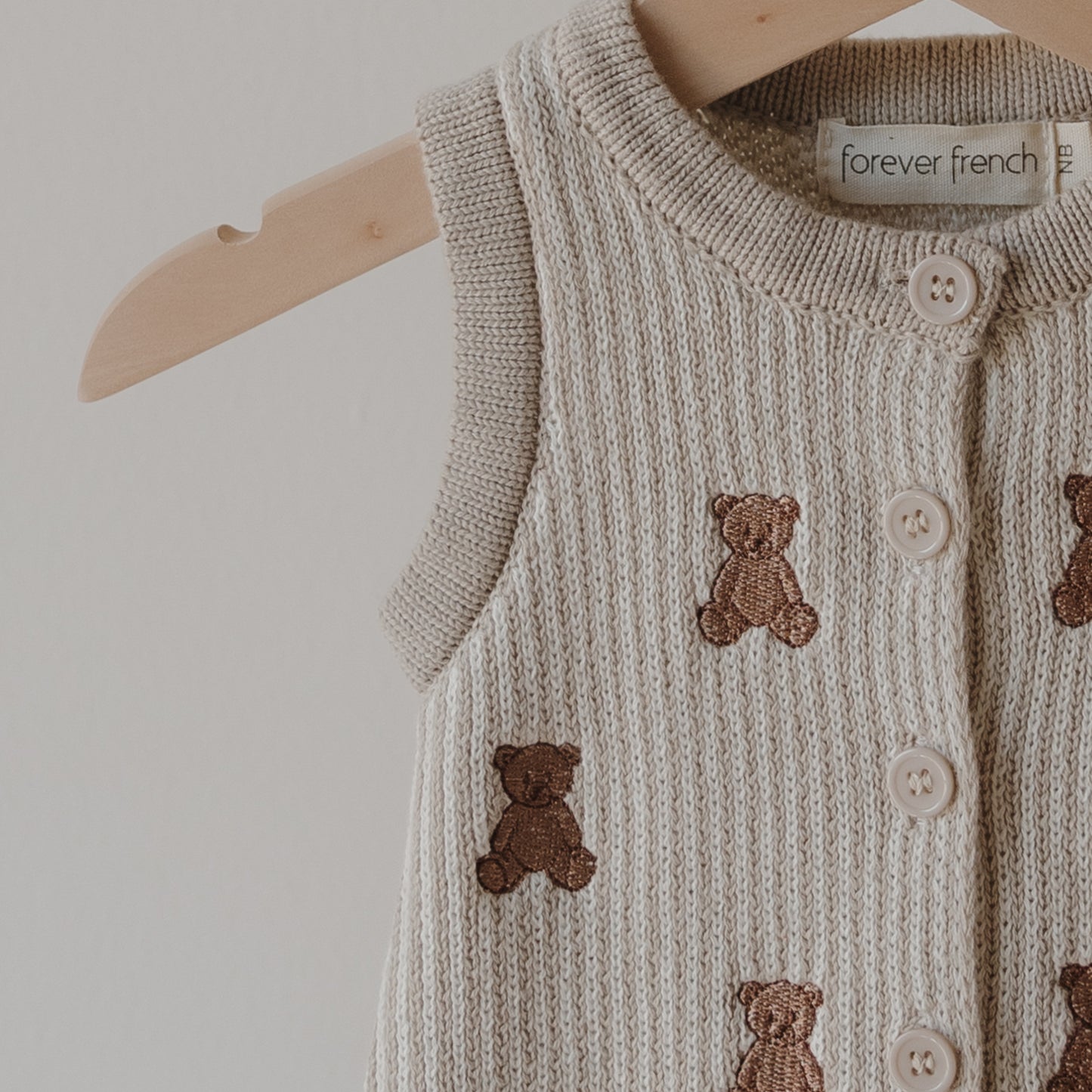 The forever french baby Knit Romper | Teddy is a beige sleeveless 100% cotton knit with white buttons and embroidered teddy bears, displayed on a wooden hanger against a light background.