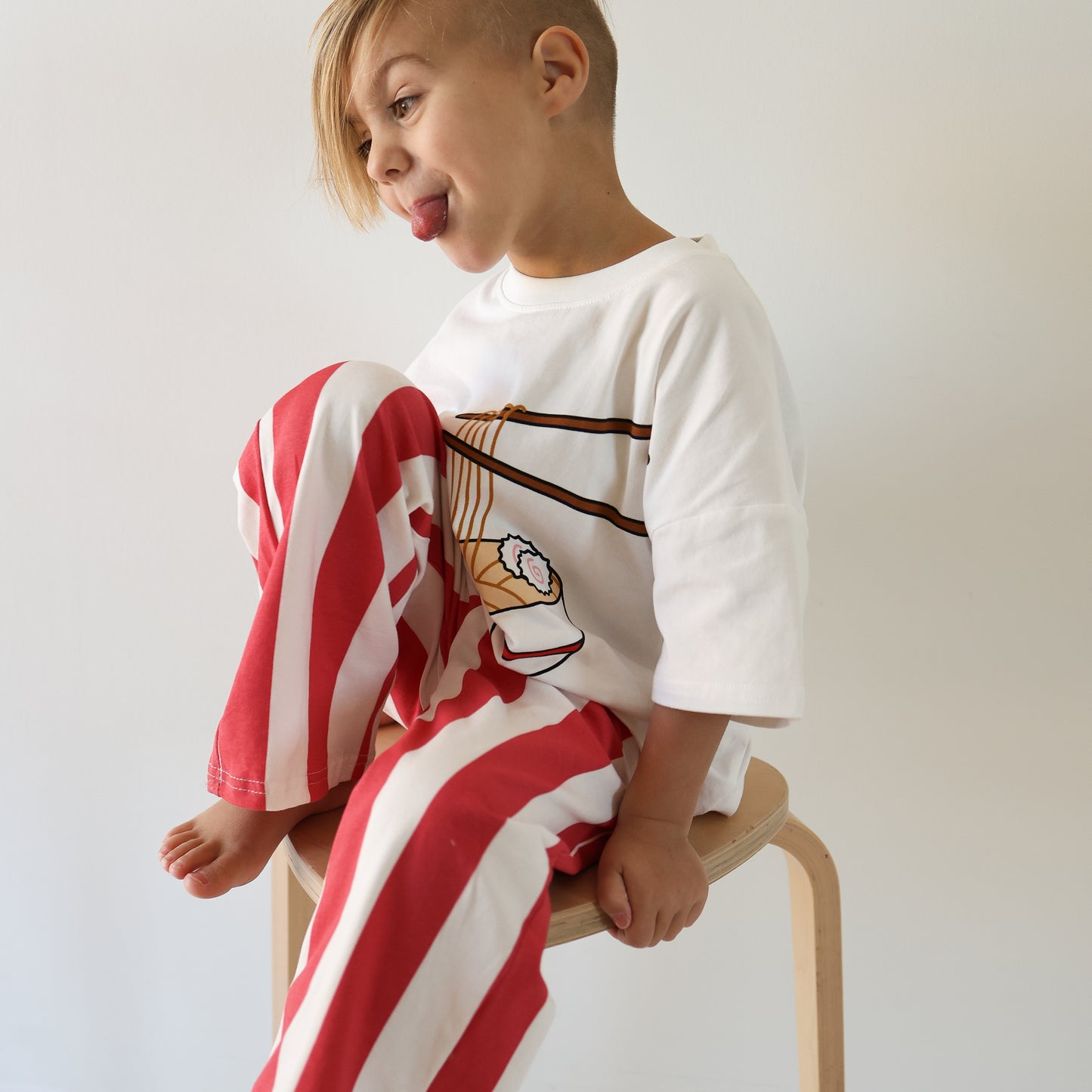 A young child with short, side-swept hair sits on a wooden stool, playfully sticking out their tongue. They wear the forever french baby T-Shirt & Pant Set | Ramen—an adorable kids’ daywear combo popping against the plain background.