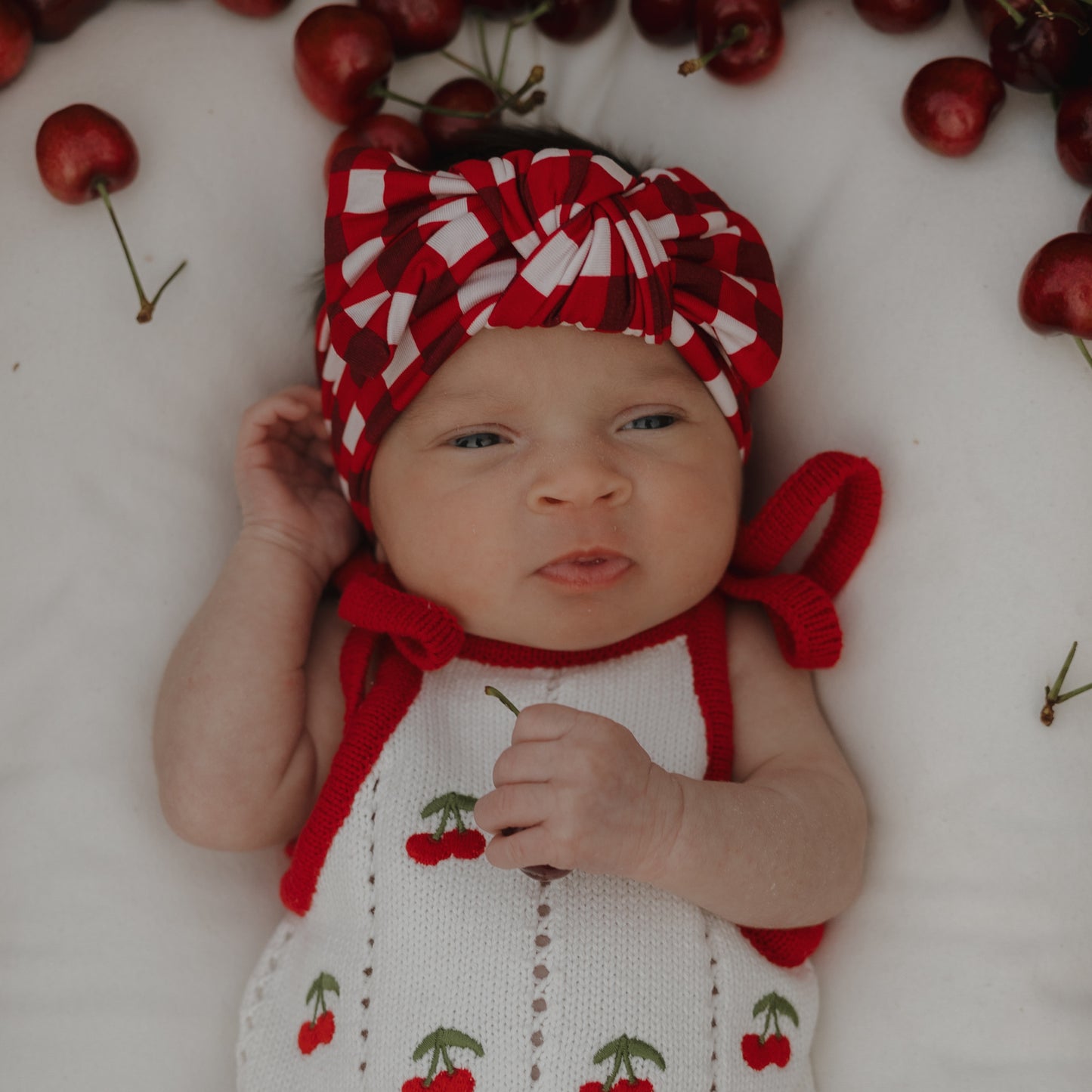 A baby wearing the forever french baby Knit Ruffle Romper | Cherry with red cherry patterns and adjustable tie straps lies on a white surface, accessorized with a red and white checkered headband and surrounded by fresh cherries.