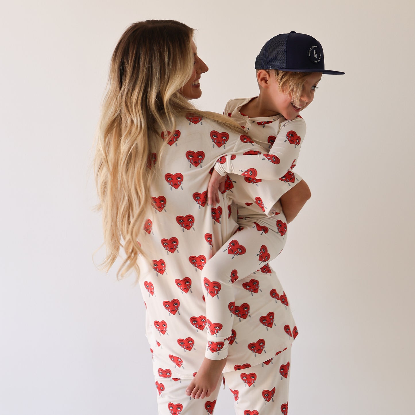 A woman with long blonde hair holds a smiling child in matching forever french baby Bamboo Two Piece Pajamas | Sad Girl, featuring red heart patterns; the child also wears a navy blue cap. They pose against a plain light background.