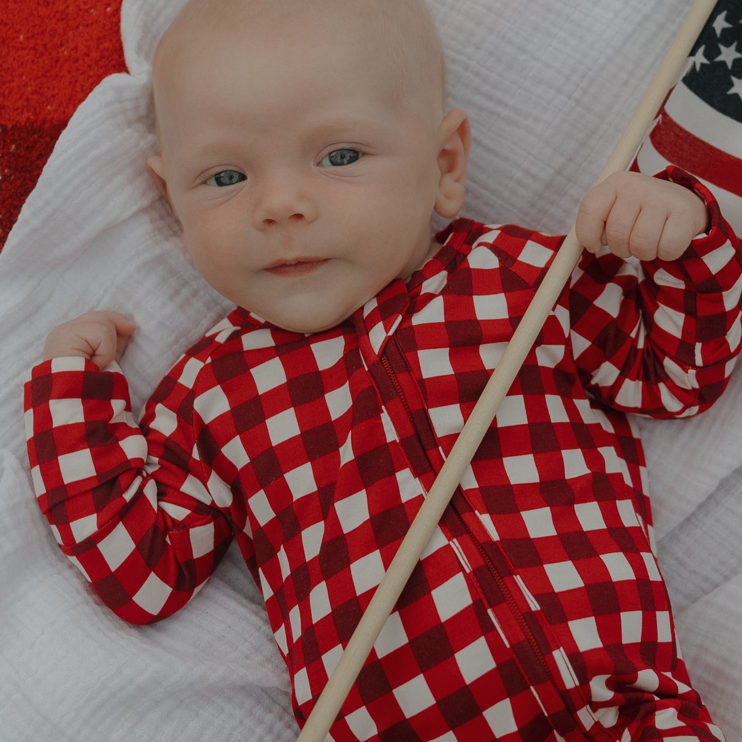 A baby wearing forever french baby Bamboo Zip Pajamas in Scarlet Gingham lies on a white blanket, holding a small American flag.