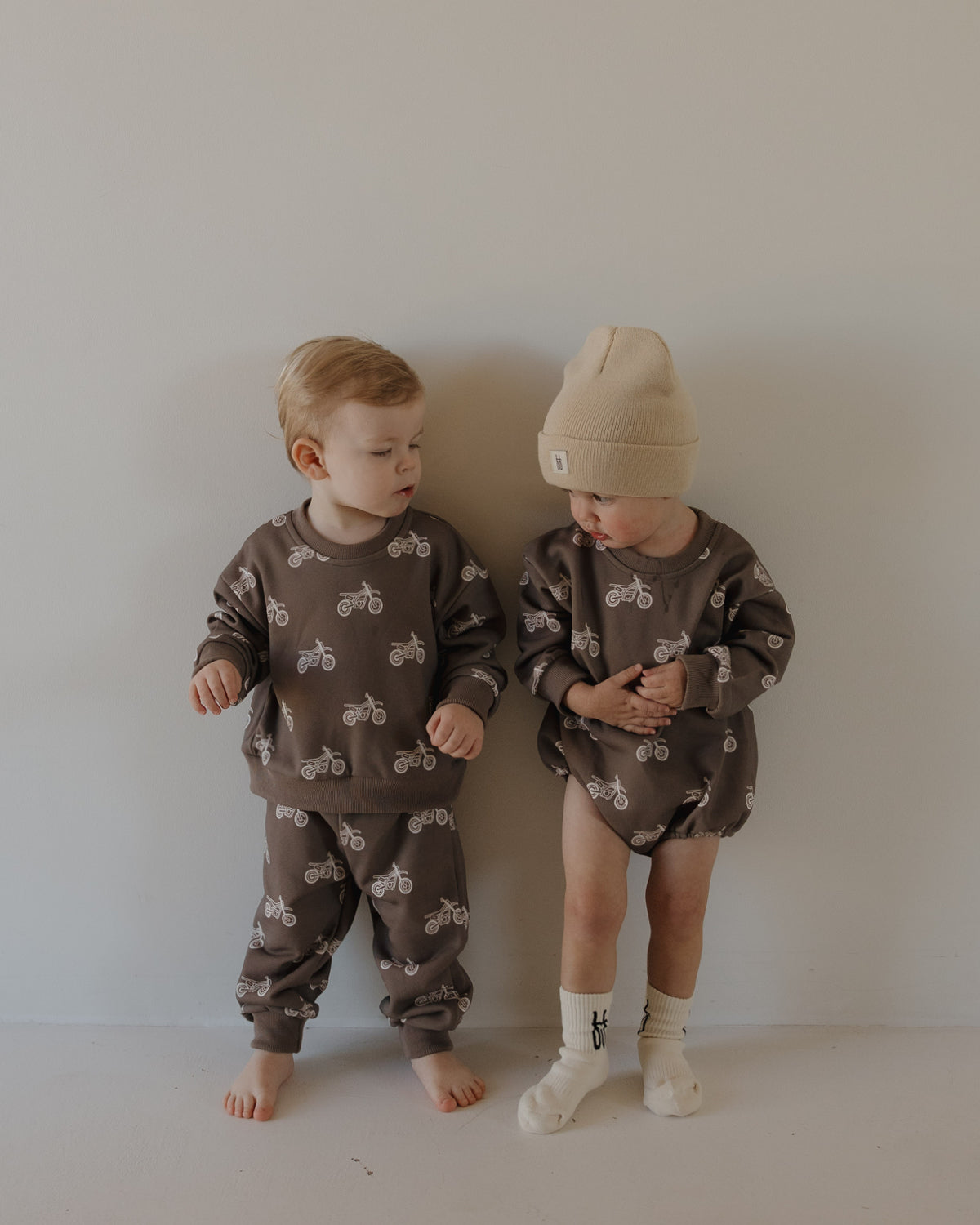 Two young children stand barefoot by a plain wall, wearing forever french baby’s Child Sweat Set | Dirt Bike—one in pants, the other in shorts and a beige beanie—both sets adorned with the playful dirt bike print.