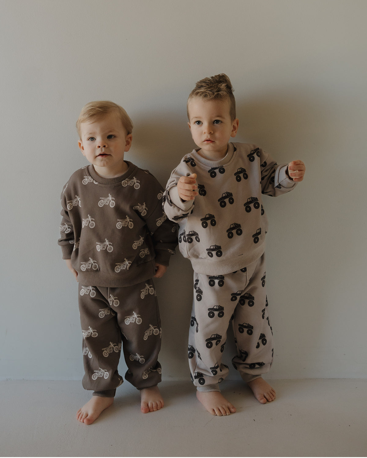 Two kids stand barefoot by a light wall, each in forever french baby's Child Sweat Set | Monster Truck—cozy long-sleeve pajamas, one in brown and one in beige, perfect for everyday adventures.