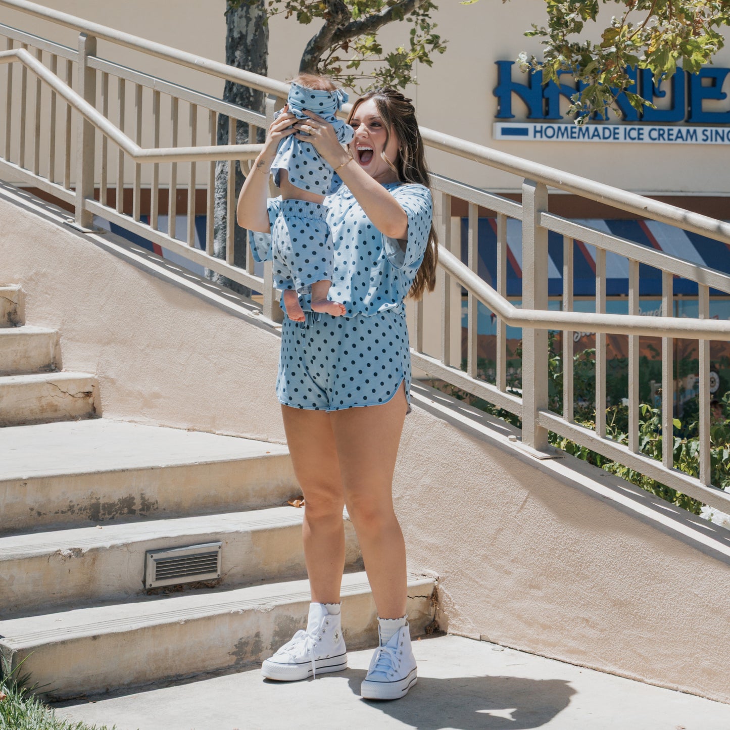 A woman in blue polka-dot clothes joyfully lifts a smiling baby, who is wearing the forever french baby Child Short Set | Sully, by some outdoor steps. Both are dressed in matching family outfits, with a homemade ice cream shop sign visible in the background.