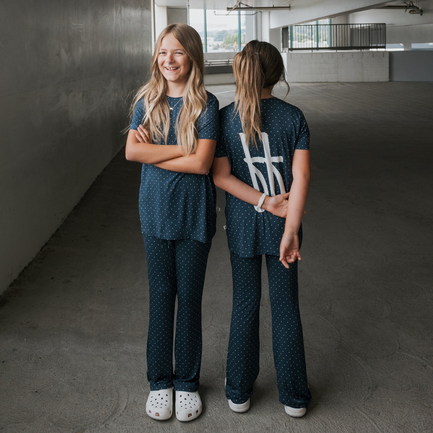 Two girls wear matching forever french baby Child Short Sleeve Bamboo Lounge Set | Emery in navy blue polka dots—one smiles with crossed arms, the other flashes a peace sign. Effortlessly stylish and breathable kids’ clothing.