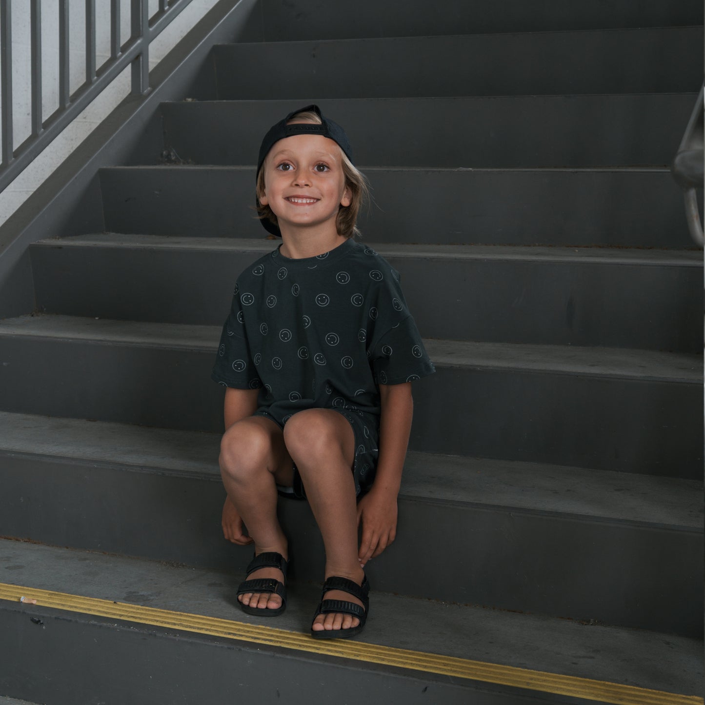 A young boy in a Child Short Set | Smiley by forever french baby and sandals smiles while sitting on gray stairs near a railing, wearing his black cap backwards and a stylish cotton blend top.