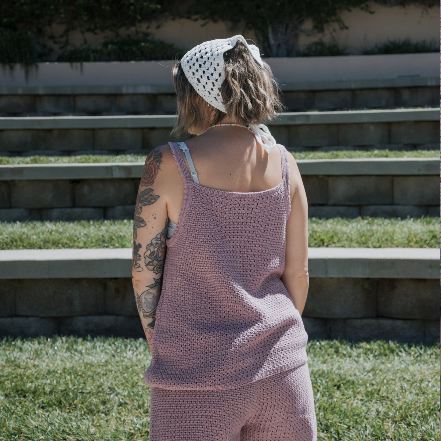 A person with short hair, visible tattoos, and a white crochet headscarf stands outdoors on grass, facing away. They are wearing the Women's Knit Short Set | Matilda by forever french baby—a sleeveless lilac matching set crafted from 100% cotton crochet. Stone steps and greenery can be seen in the background.