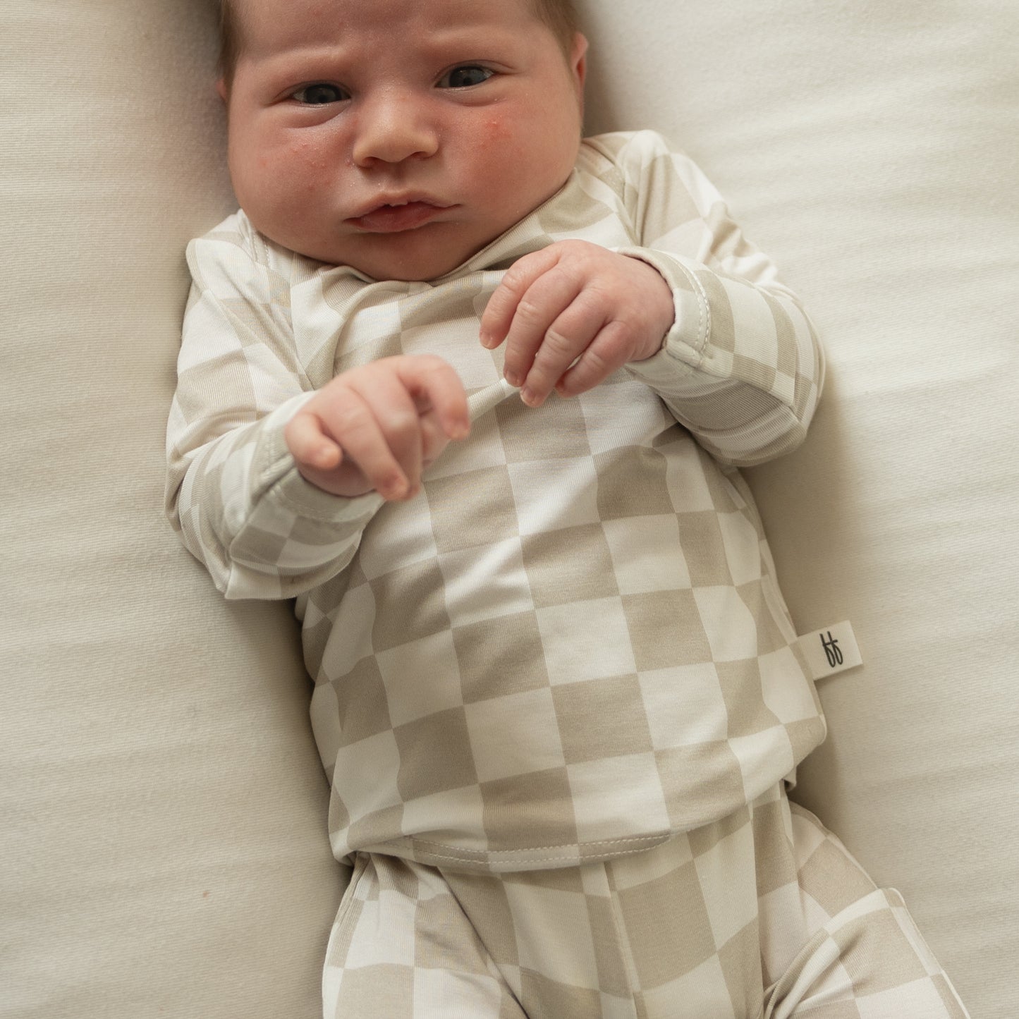A newborn rests on a cream blanket, wearing the forever french baby Sleepy Time Set | Checkerboard in beige and white, gazing up calmly with gently clenched fists.