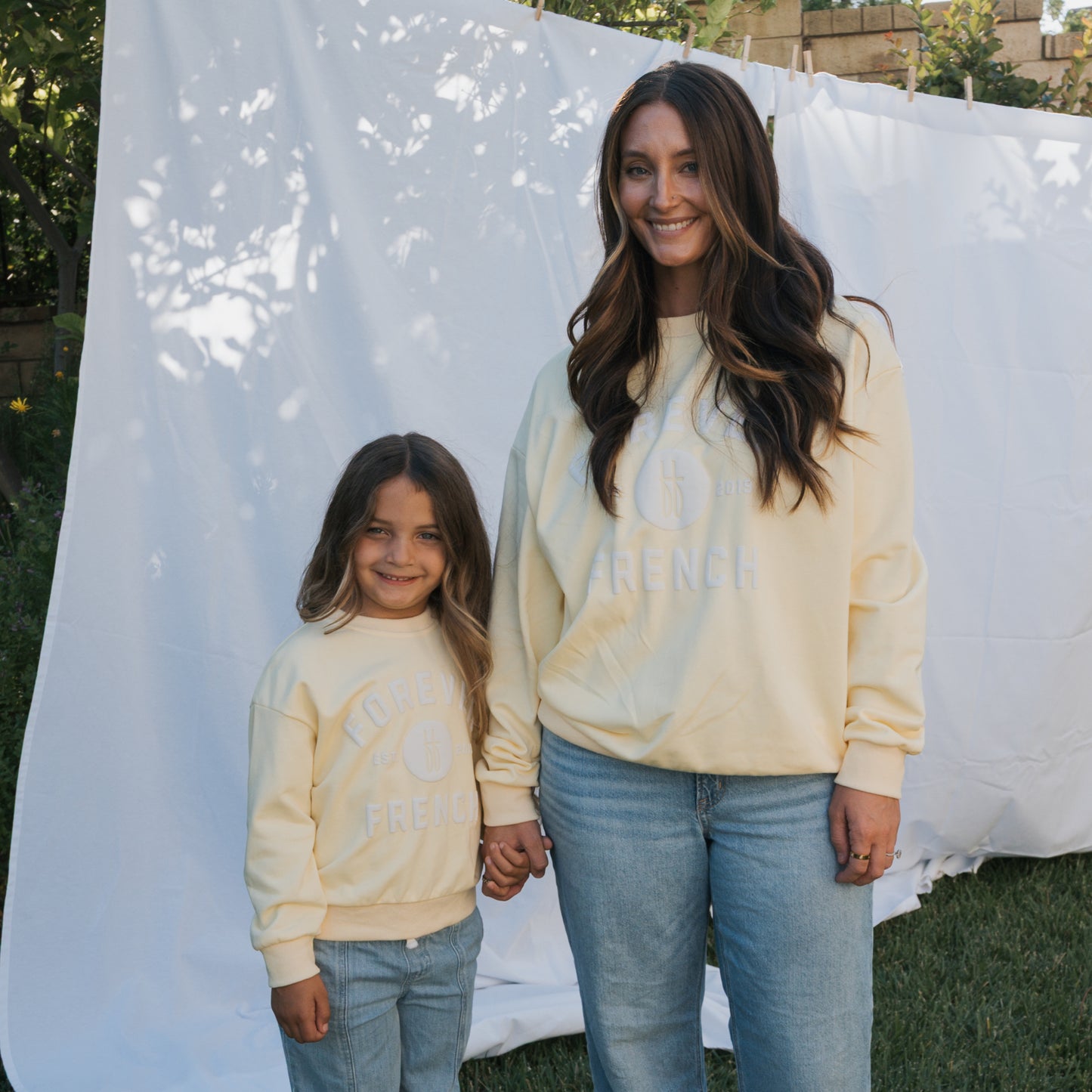 A woman and a young girl, both smiling and holding hands outdoors, wear matching blue jeans and forever french baby Child Sweatshirt | ff Yellow—light yellow 100% cotton sweatshirts—in front of a white backdrop.