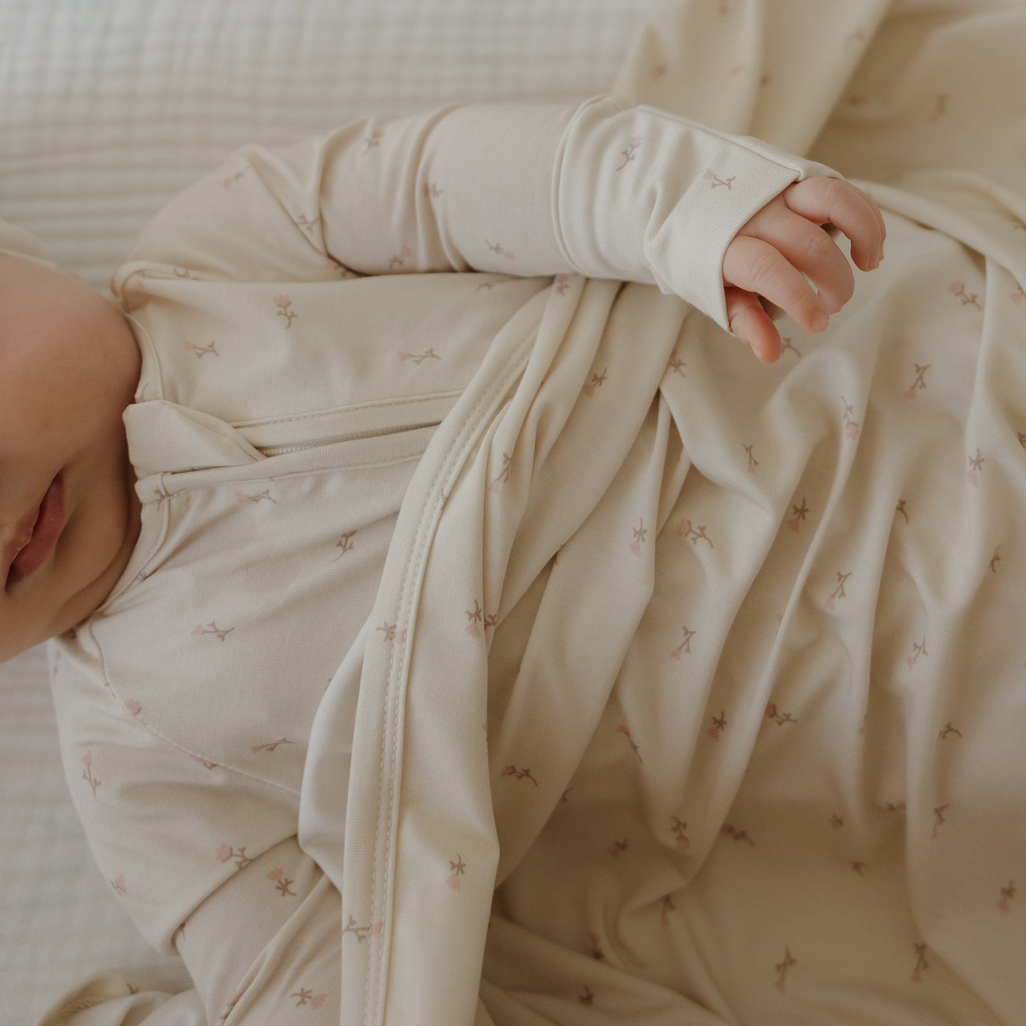 A baby in a soft, cream-colored onesie with star patterns lies on the forever french baby Bamboo Swaddle | Lucy. Only part of the baby's face and one arm are visible.