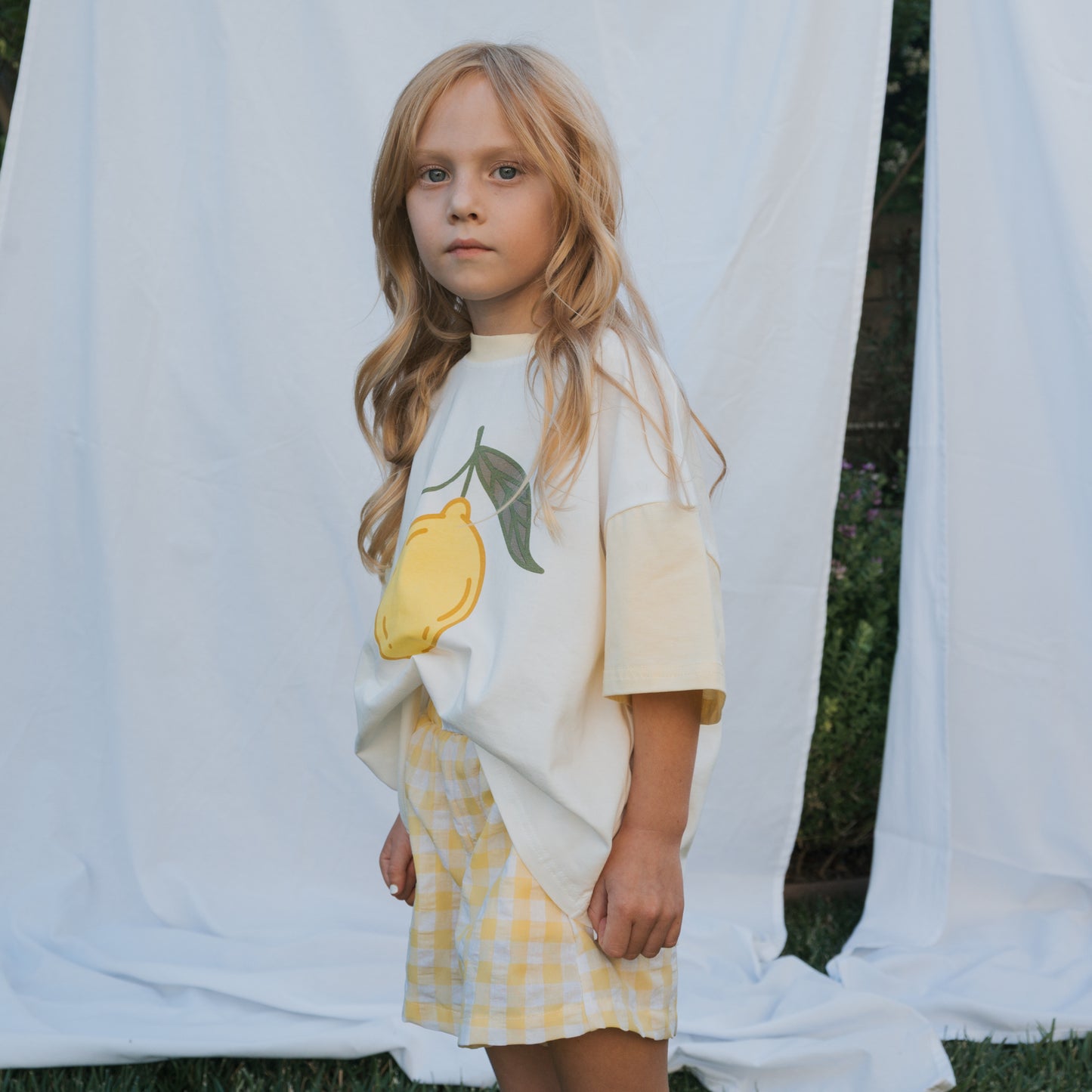 A young girl with long blonde hair stands outside before a white sheet, calmly gazing at the camera in a forever french baby Child Cotton Tee | Lemon and yellow gingham shorts—perfect for summer.