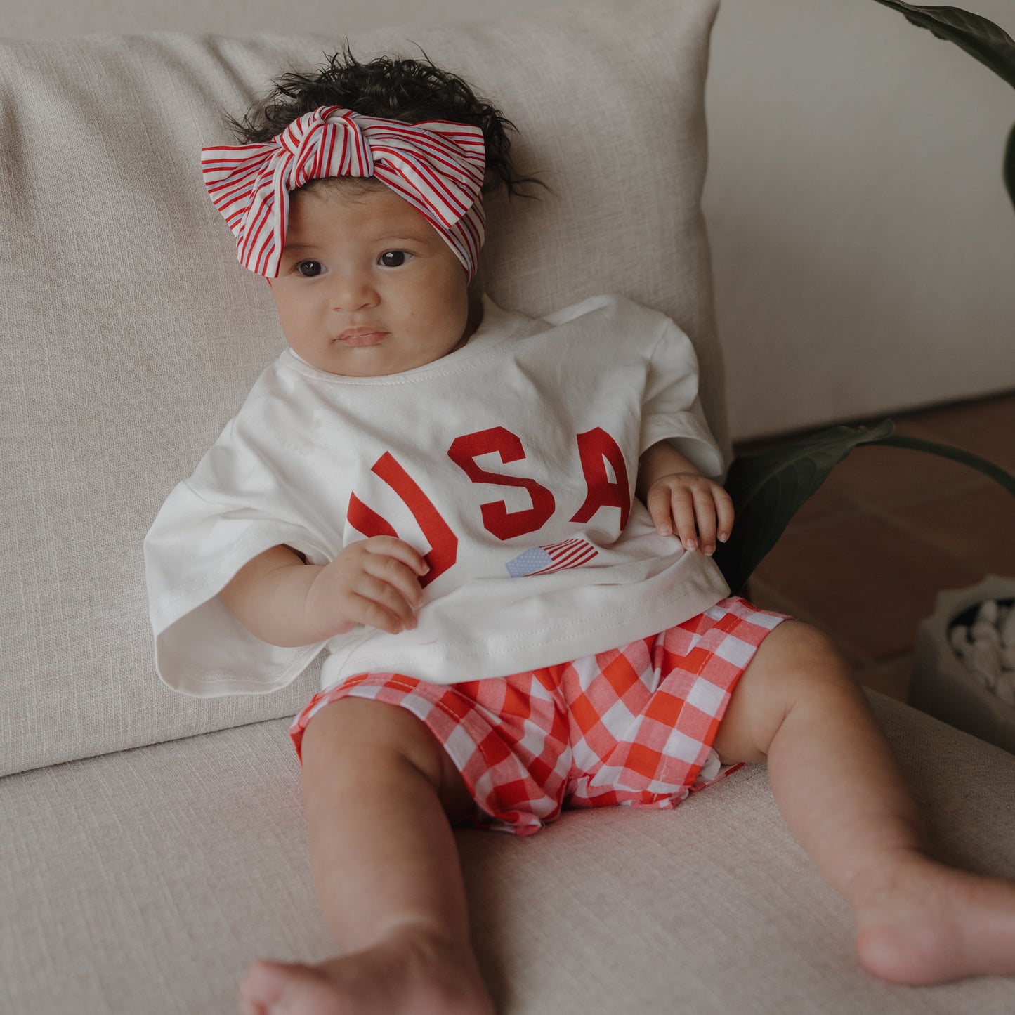 A baby sits on a beige couch wearing a white "USA" shirt, red-and-white checkered shorts, and the Bamboo Head Wrap | Heritage Stripe by forever french baby, gazing slightly to the side with a neutral expression.